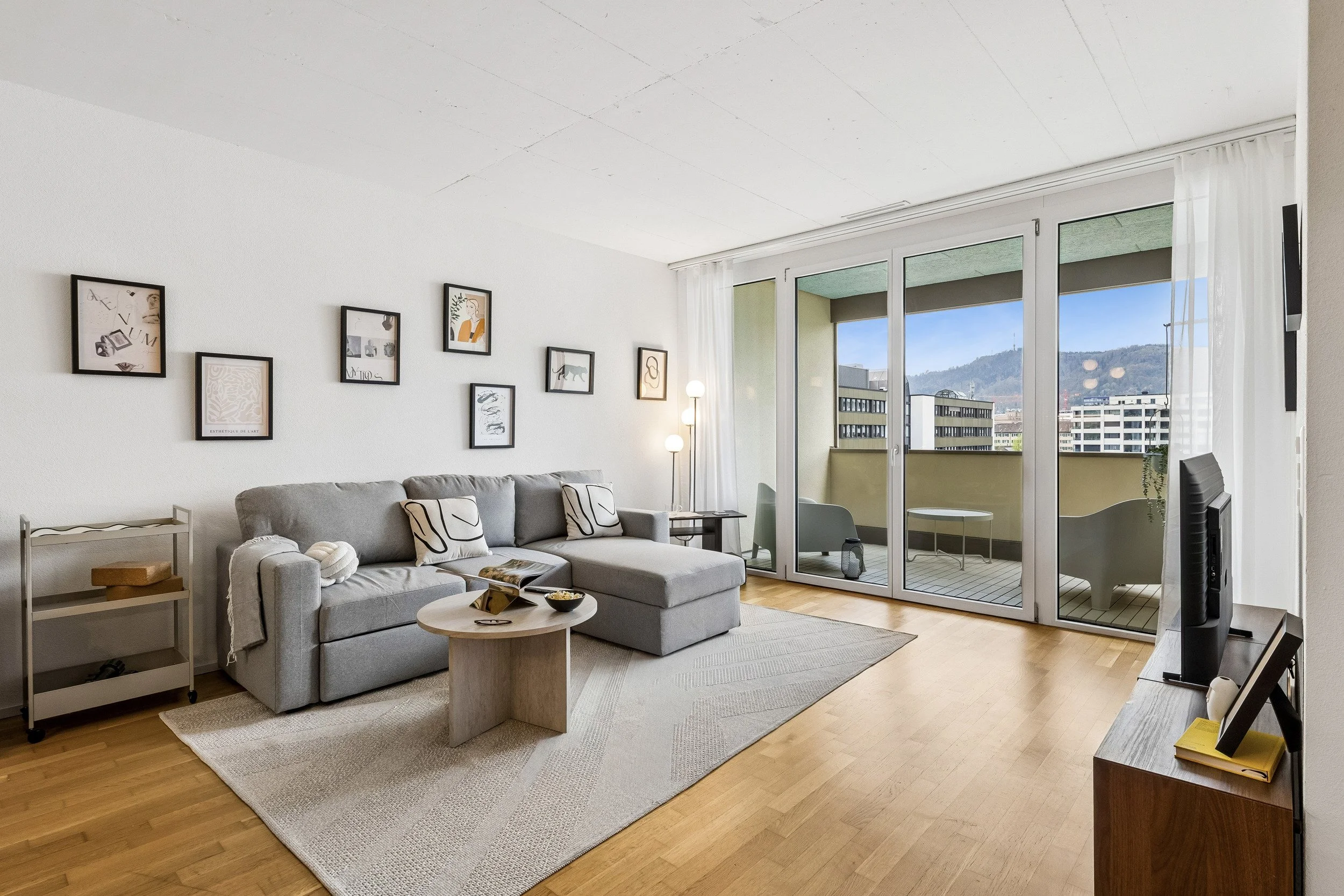 Living room with a gray sofa, framed artwork on white wall, coffee table with snacks, and large glass doors leading to a balcony with a view of city buildings and mountains.