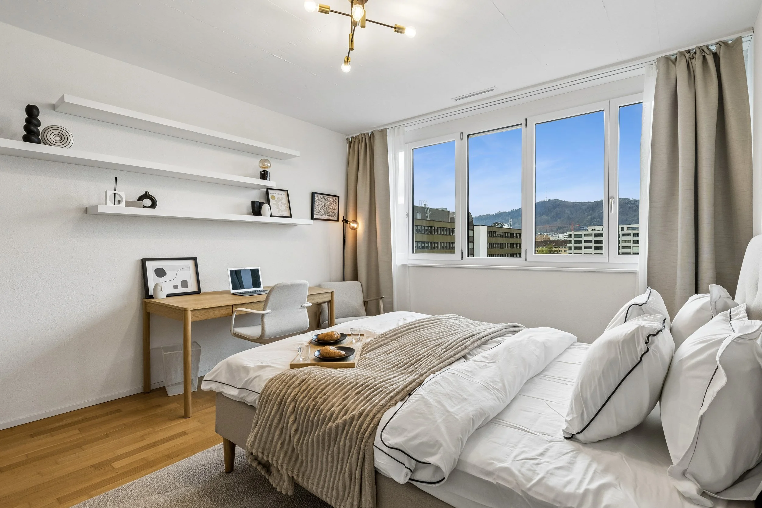 A modern bedroom with a large window showing a mountain view. The room features a neatly made bed with white bedding and black piping, beige throw blanket, and pillows. A simple wooden desk with a laptop, framed artwork, and a white chair are beside 