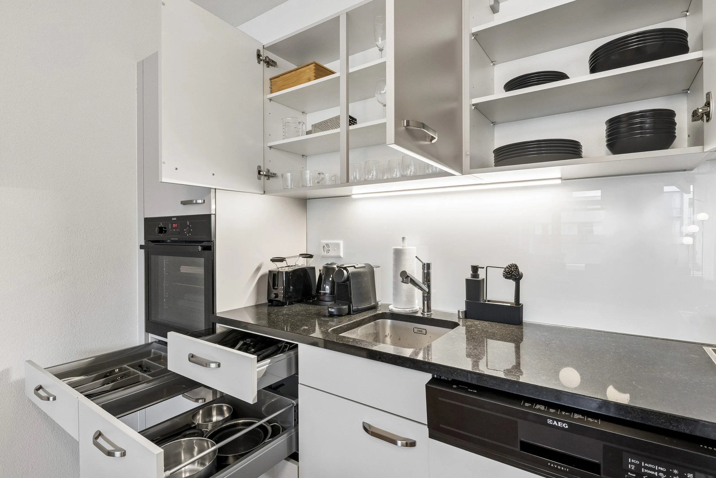 Modern kitchen with open cabinets, black countertop, and various appliances including a built-in oven, toaster, and coffee machine. Open drawers reveal utensils and kitchen tools.