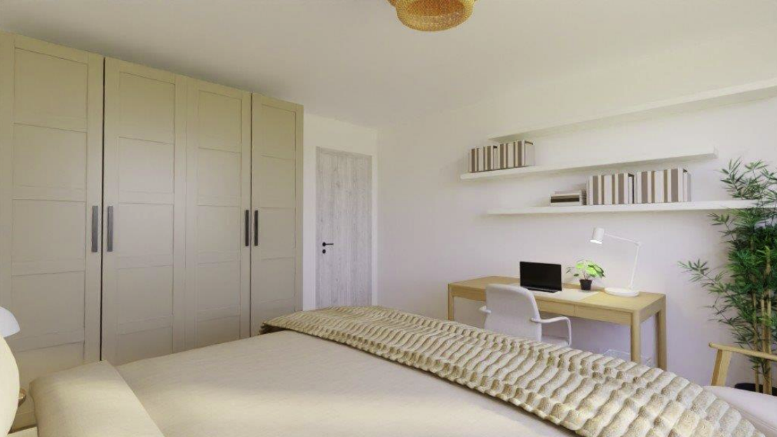 Bedroom with beige wardrobe, bed, desk with lamp and laptop, white chair, floating shelves with books, and green plant.