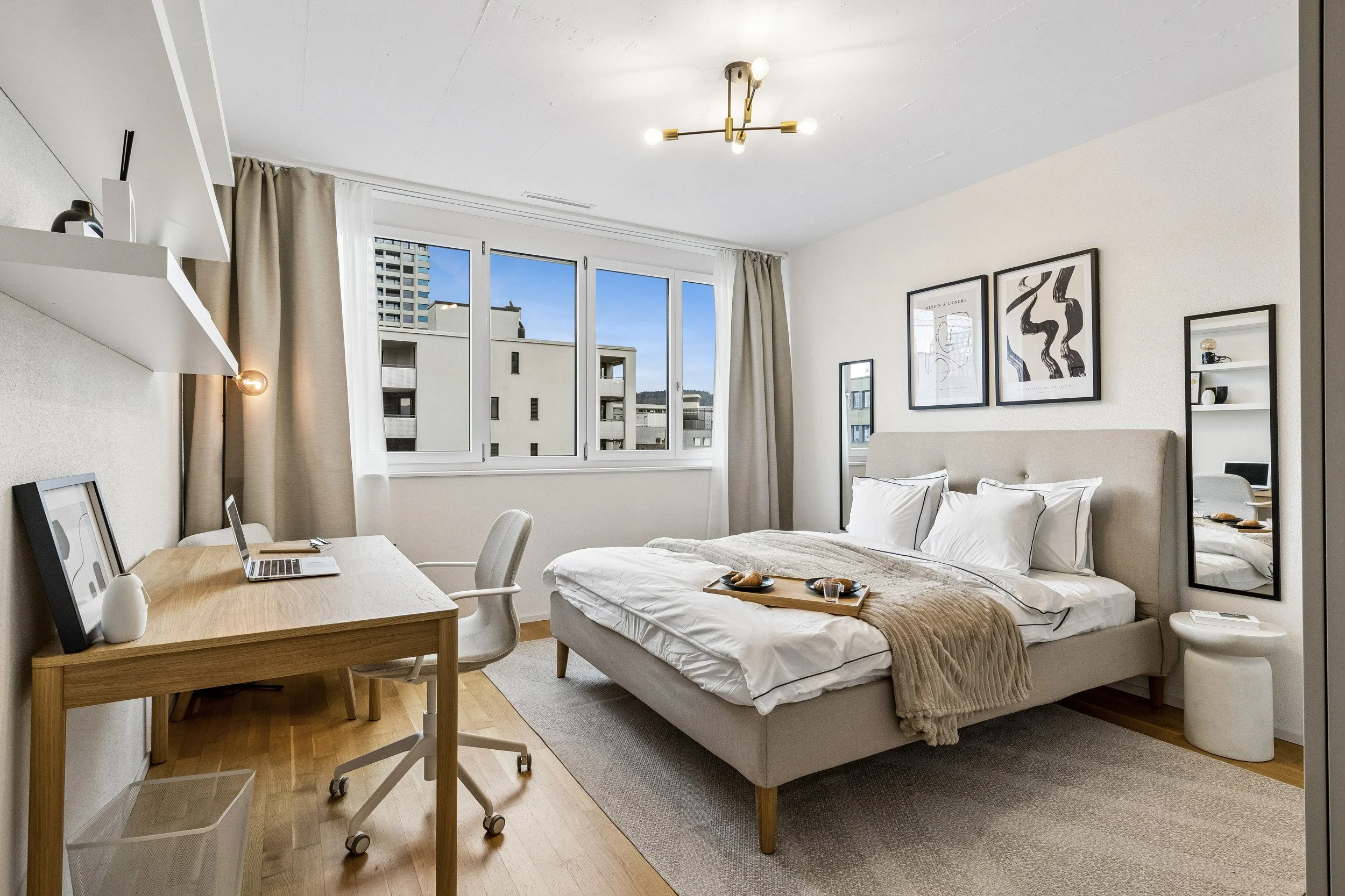 A bedroom with a large window showing neighboring buildings, a bed with white linens and beige blanket, framed abstract art on the wall, a wooden desk with a laptop, and modern lighting fixtures.