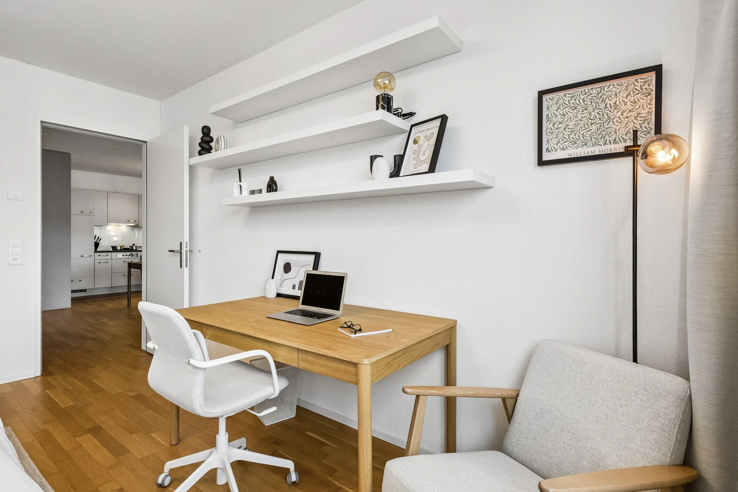 A modern home office with a wooden desk, white ergonomic chair, and beige armchair. White floating shelves with decorative items are mounted on the wall, and a framed botanical print is hanging above a floor lamp with an amber glass shade. The floor 