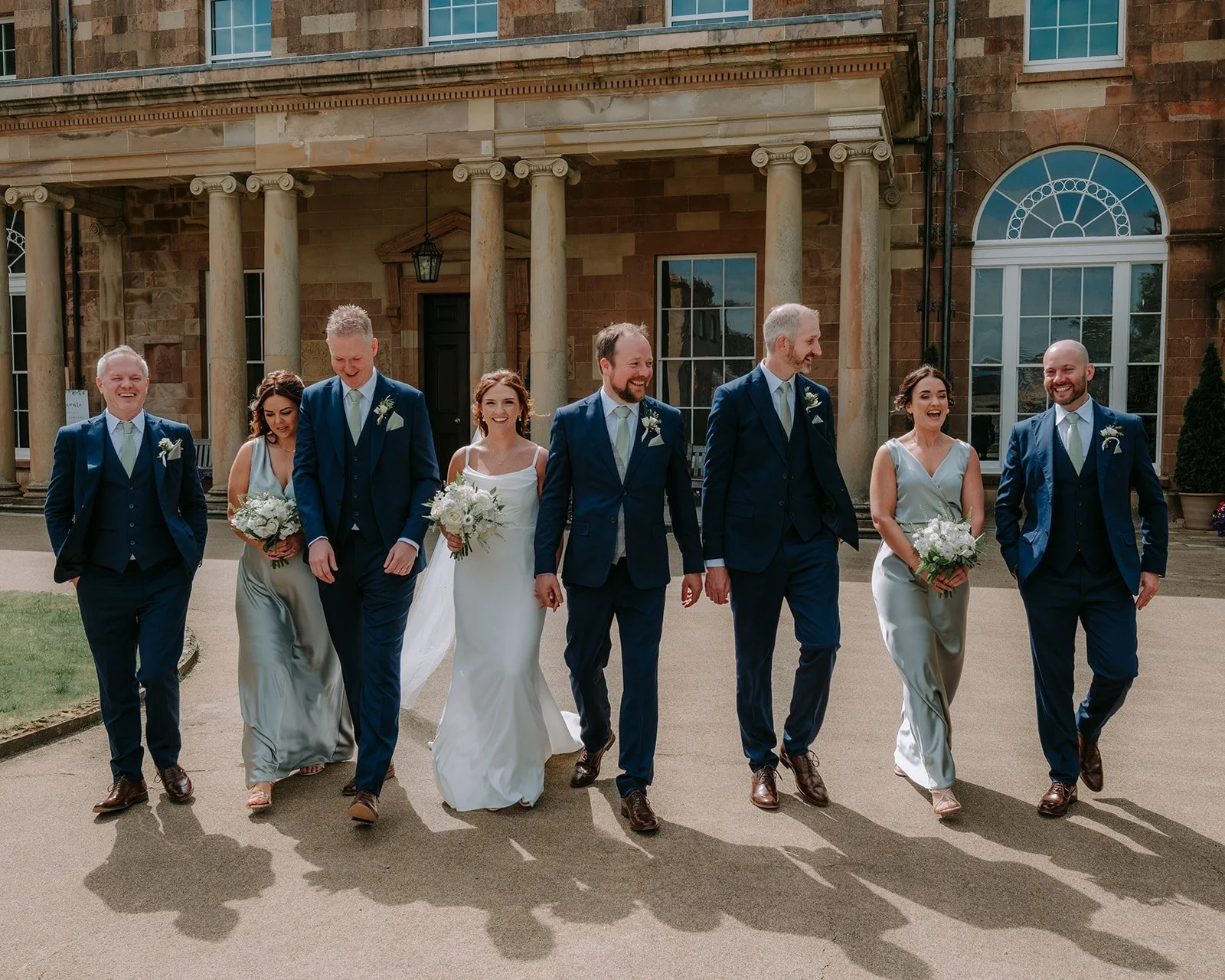 Wedding party walking together, with bridesmaids in silver dresses and groomsmen in blue suits, in front of a historic building with columns.
