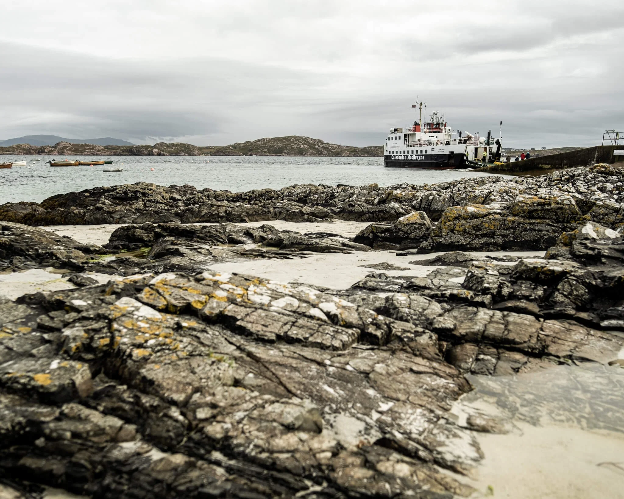 Rocky beach with boats and a ferry in the distance, under cloudy sky.