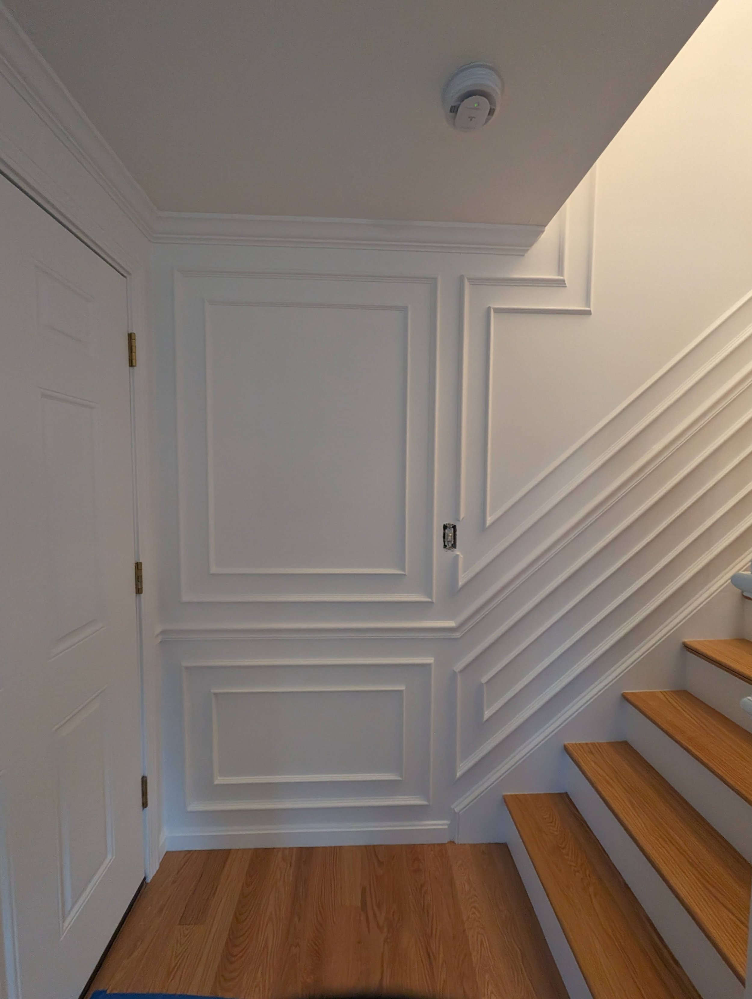 Interior view of a staircase with white paneled walls, a ceiling smoke detector, a door on the left, and wooden stairs on the right.