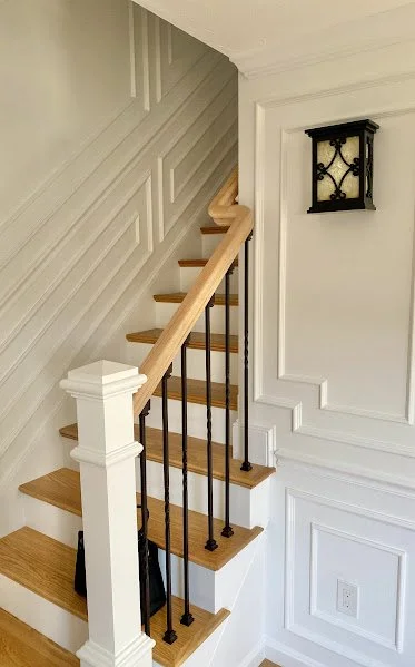 Interior view of a staircase with wooden steps, a wooden handrail, black metal balusters, decorative white wall paneling, and a wall-mounted black lantern-style light fixture.