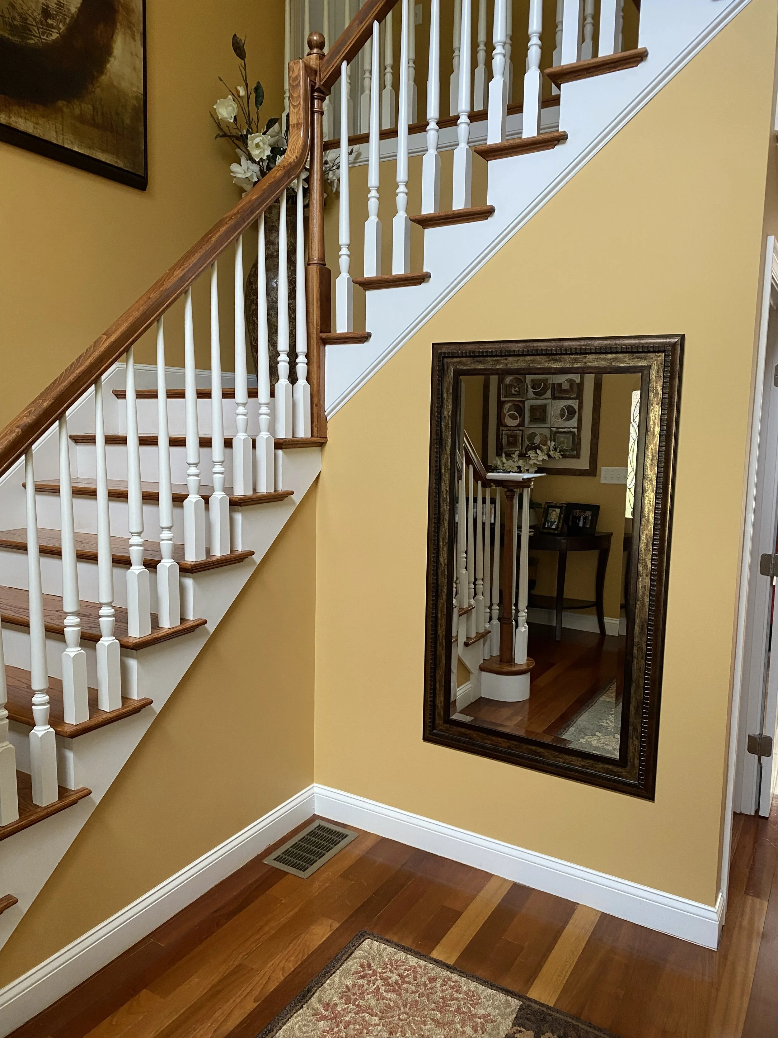 A staircase with wooden steps and white spindles, a large mirror on a yellow wall, a decorative flower arrangement, and framed artwork. The room has hardwood floors and a vent on the floor.