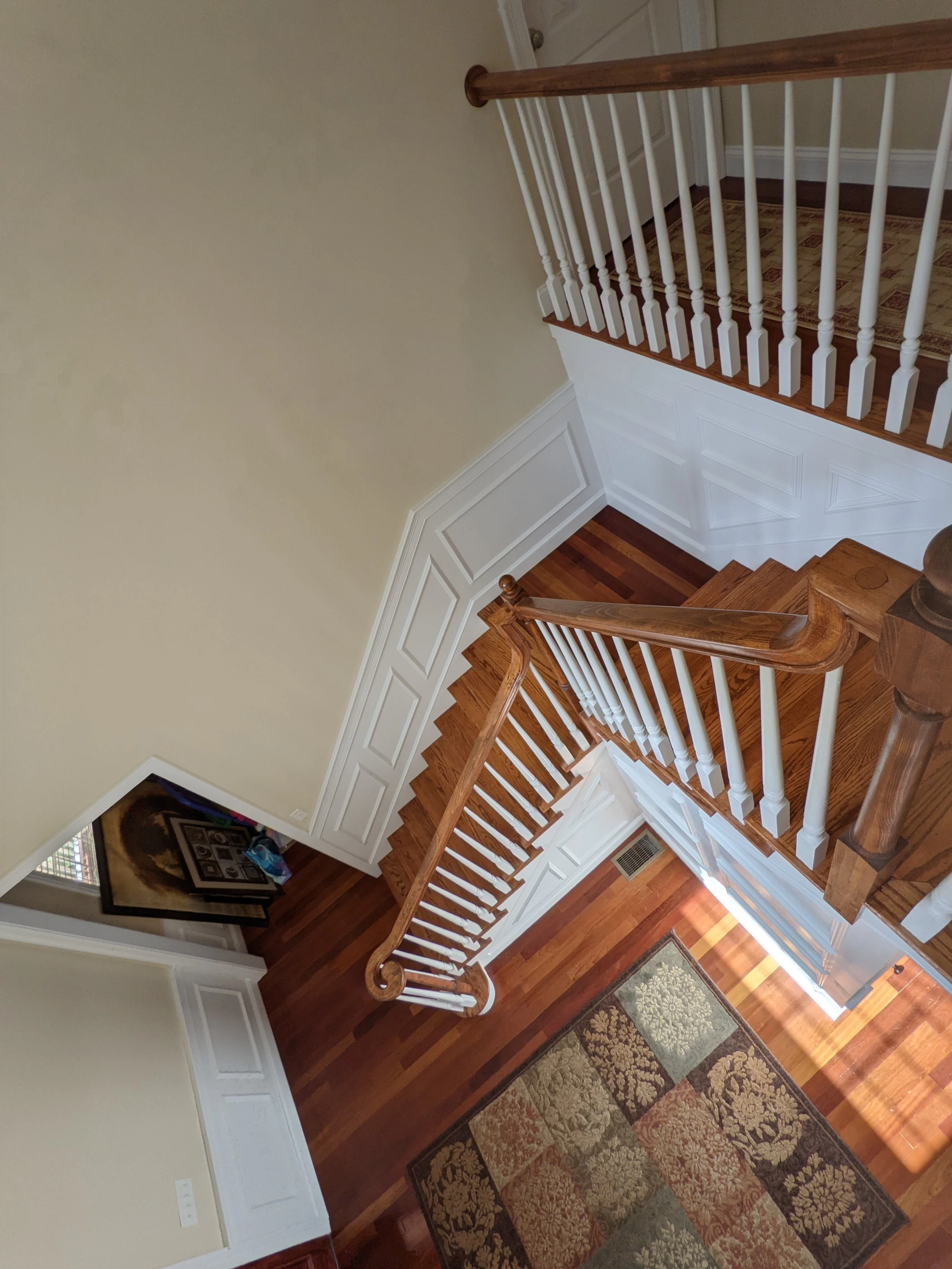 View of a staircase with wooden steps and white balusters, leading to a hallway with hardwood flooring, a decorative rug, and a wall with paneling and a door.