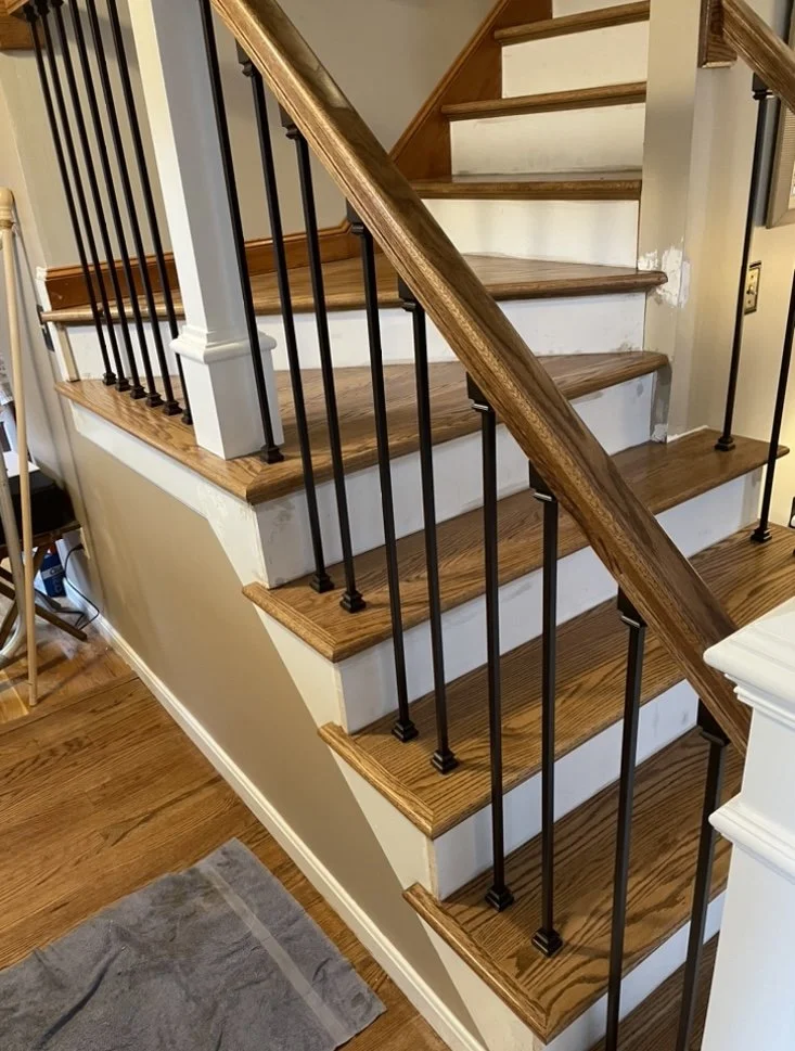 Interior view of a wooden staircase with dark iron balusters, wooden handrail, and steps with visible wood grain; part of the adjacent wall, baseboard, and a gray rug are also visible.