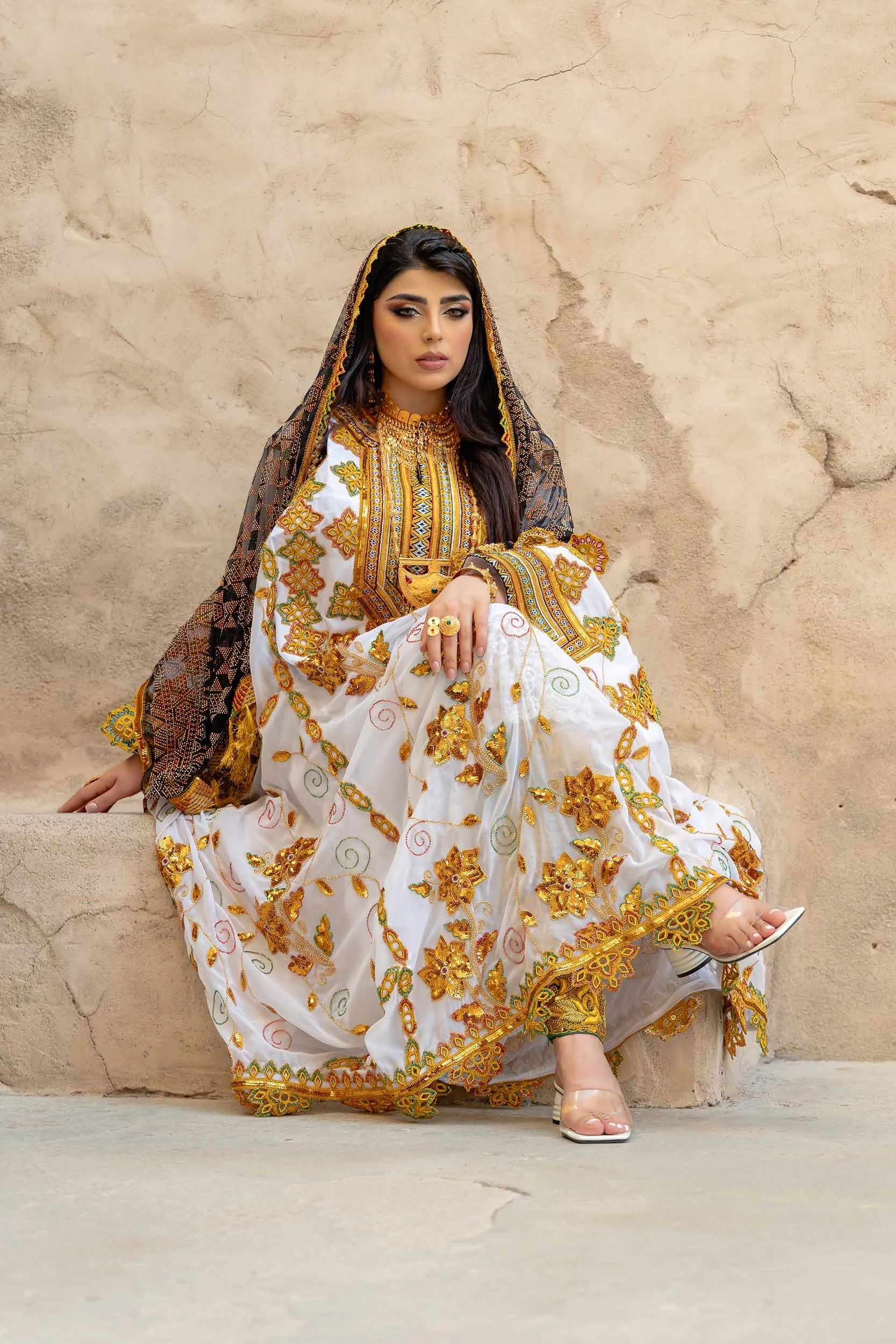 A woman dressed in traditional, ornate white and gold embroidered attire, sitting against a beige, textured wall, wearing jewelry and high-heeled sandals.