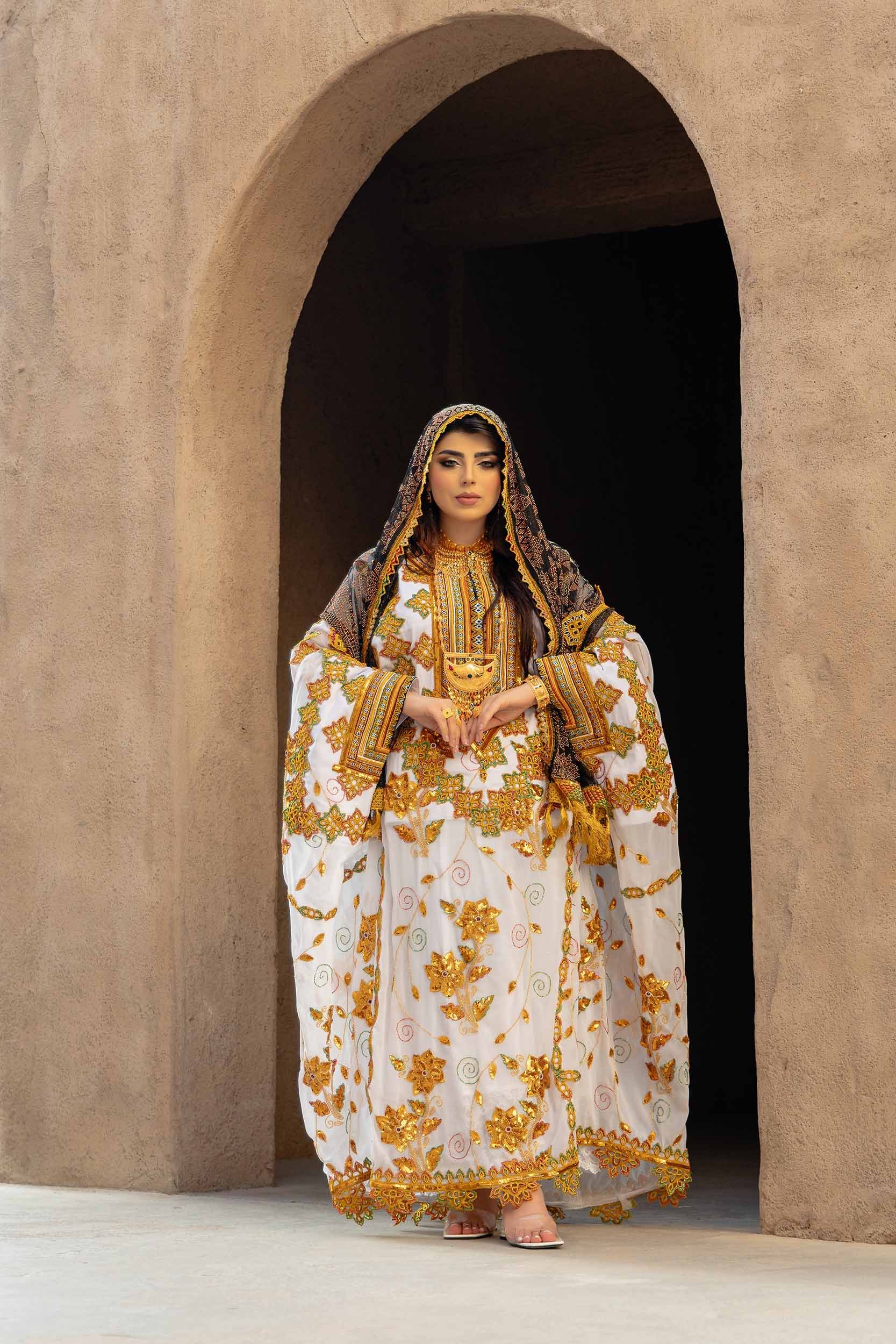 A woman dressed in traditional, ornate, embroidered white and gold attire with a matching headscarf, holding a gold bowl, standing in front of a beige textured archway.