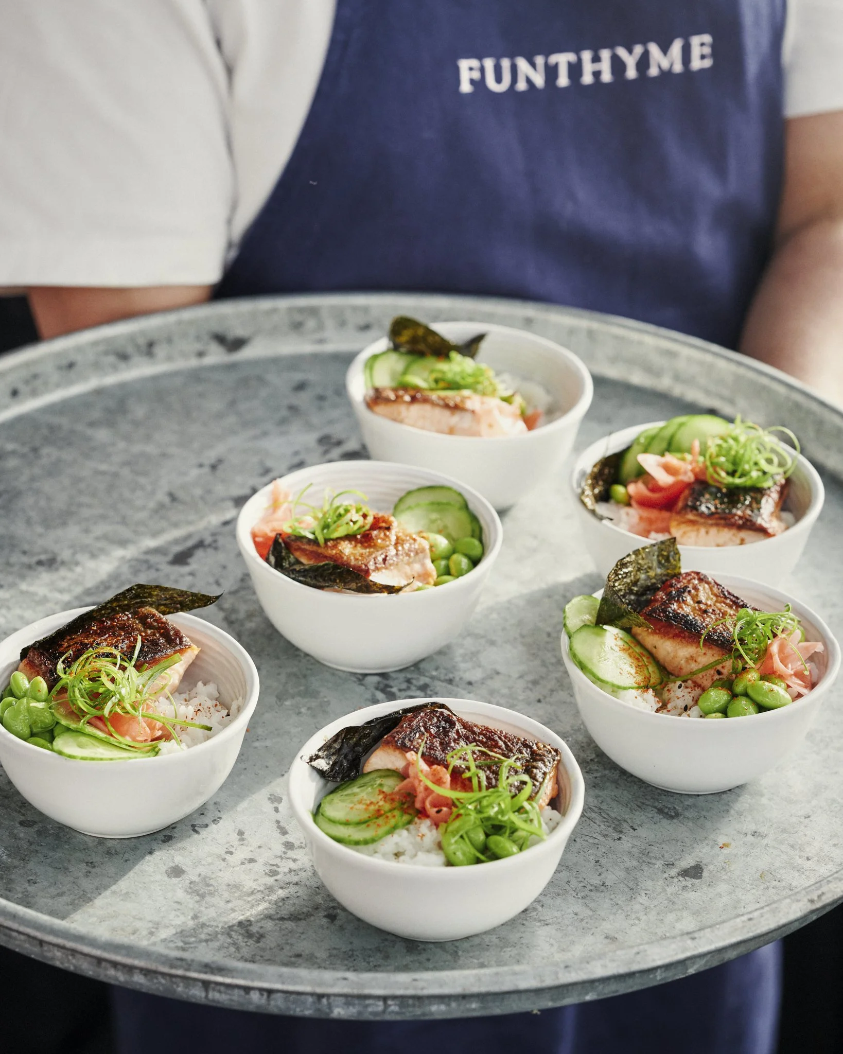 Five bowls of rice topped with glazed salmon, cucumber slices, seaweed, edamame, and green onions, served on a metal tray. The person holding the tray is wearing a blue apron with 'FUNTHYME' printed on it.