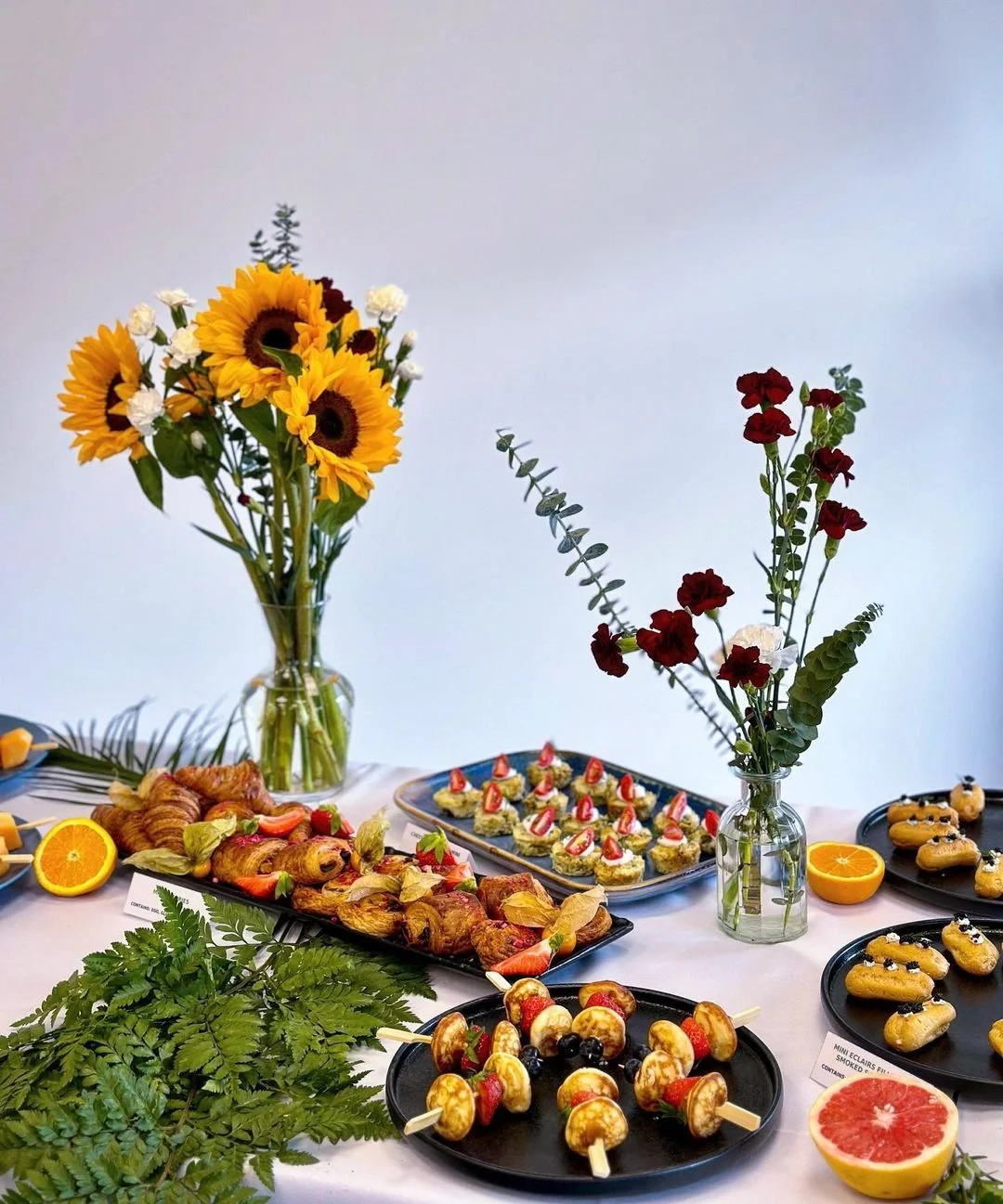 A table set with assorted pastries and skewers featuring fruit and baked goods, adorned with yellow sunflowers and red flowers in glass vases for decoration. Plates contain desserts like éclairs and bite-sized treats, surrounded by halved oranges and greenery.