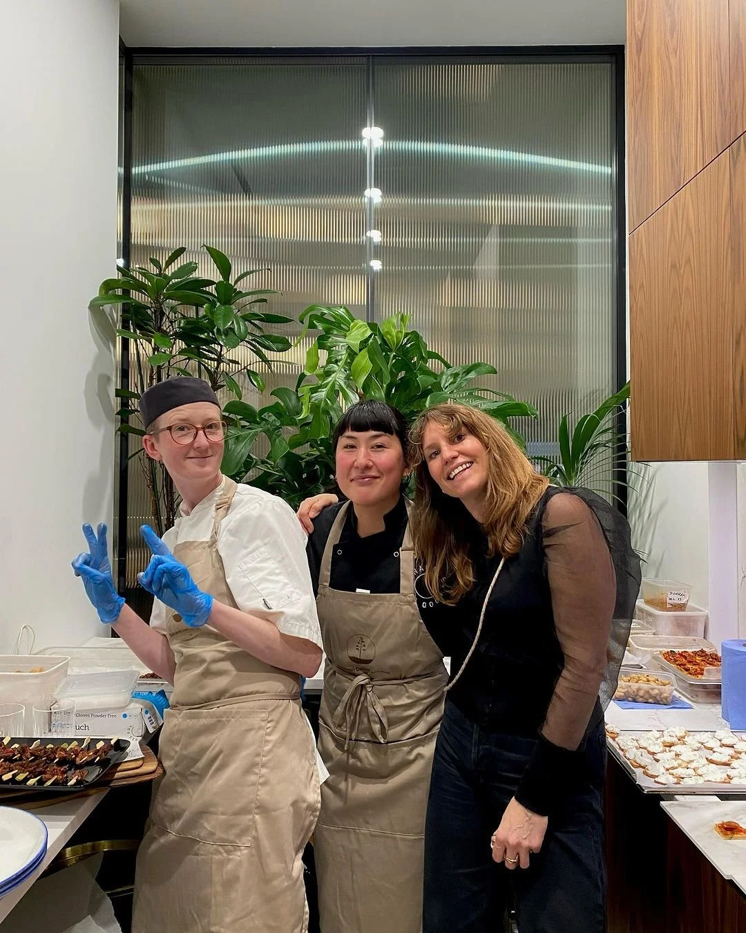 Three people posing in a kitchen setting, two wearing aprons, one in a chef's uniform with a hat and gloves, standing near a counter with food items. There are green plants in the background.