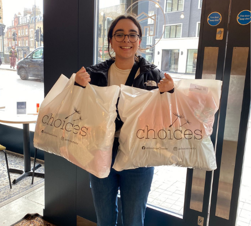 Person smiling and holding two "choices" branded shopping bags indoors near a window with a street view.