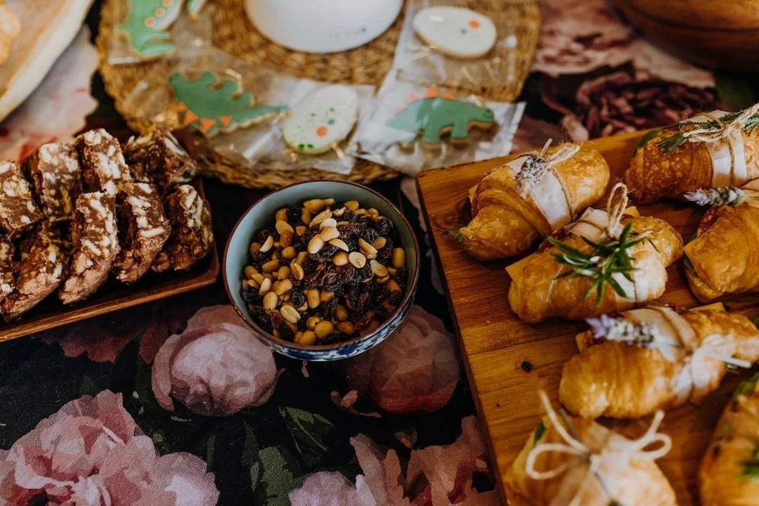 Assorted pastries and snacks on a table, including chocolate biscotti, pine nuts in a bowl, decorated sugar cookies, and croissants with rosemary.