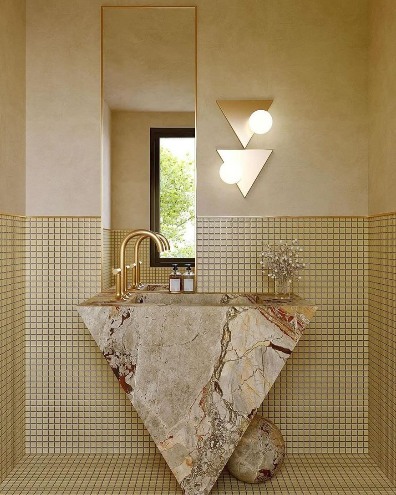 This monochromatic champagne gold bathroom is a masterpiece of impeccable style. From the geometric triangular marble sink base to the marble sphere at the bottom, every detail exudes sophistication. The triangle sconces with globe lights add a touch