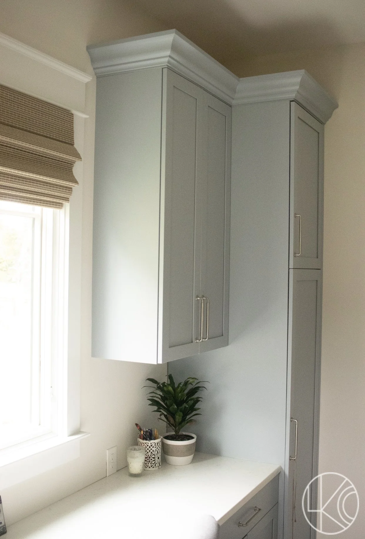 Kitchen corner with light gray cabinets, a white countertop, a potted plant, a candle, and a patterned container with pens near a window with a brown Roman shade.