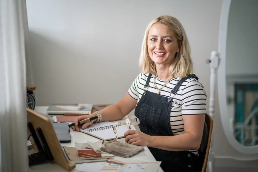 A woman with blonde hair wearing a striped shirt and black overalls sitting at a desk smiling at the camera, surrounded by design materials and tools.