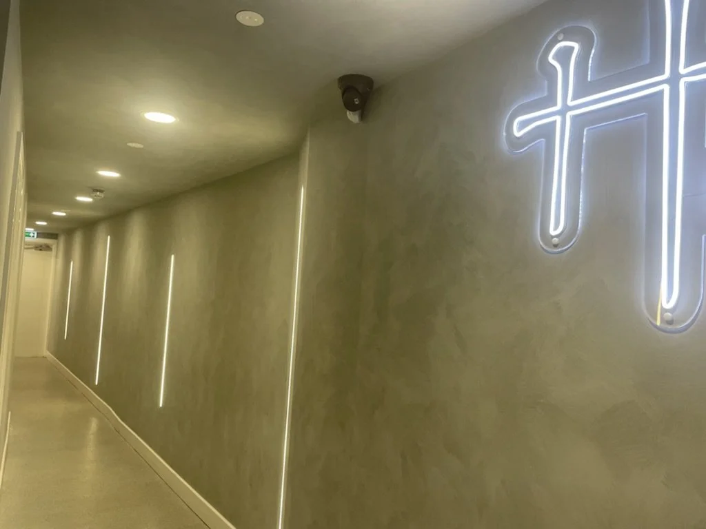 A hallway with beige walls and ceiling, illuminated by round ceiling lights and vertical LED strip lights along the wall. There is a neon cross sign on the right side of the wall.