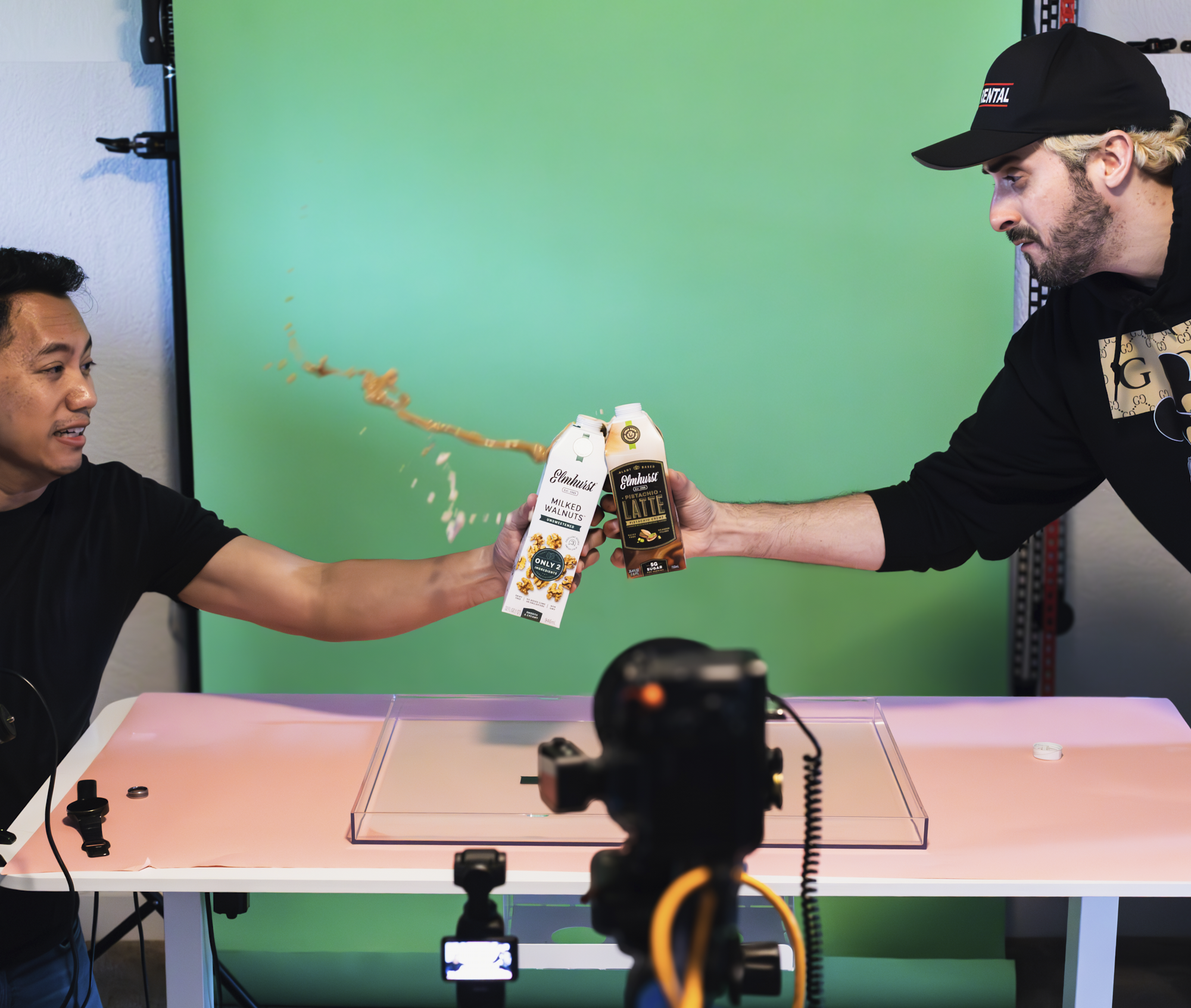 Two men seated at a table in front of a green screen, holding cartons of Elmhurst milk and coffee, with camera equipment set up in front of them and overlapping bottles of coffee on the table, creating a video recording setting.