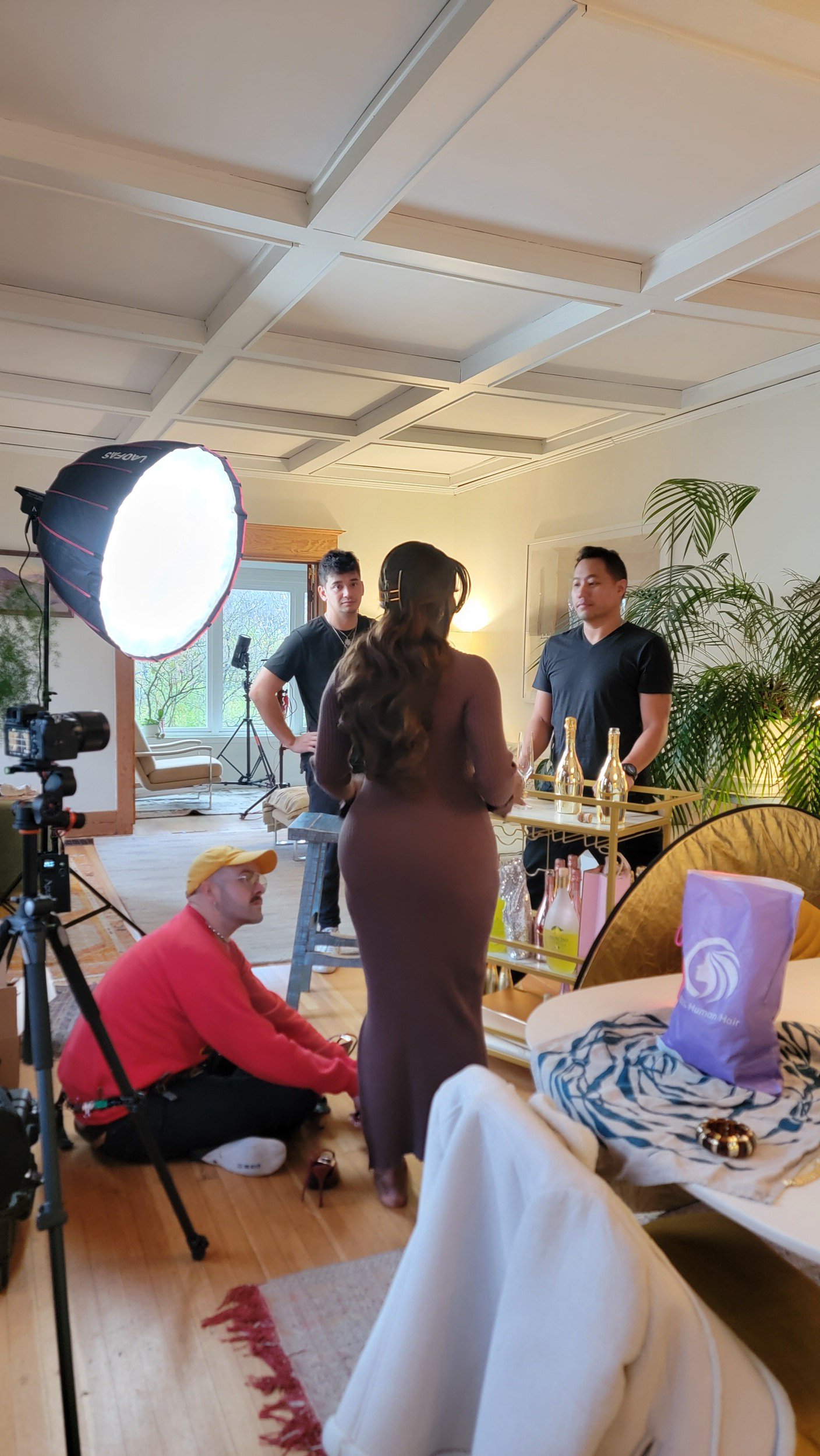 A woman in a fitted dress stands at a bar table while a man and a woman behind the bar prepare drinks. A person in a red shirt with a yellow cap is sitting on the floor. There is a large softbox light, camera equipment, and a laptop with a window showing greenery outside.
