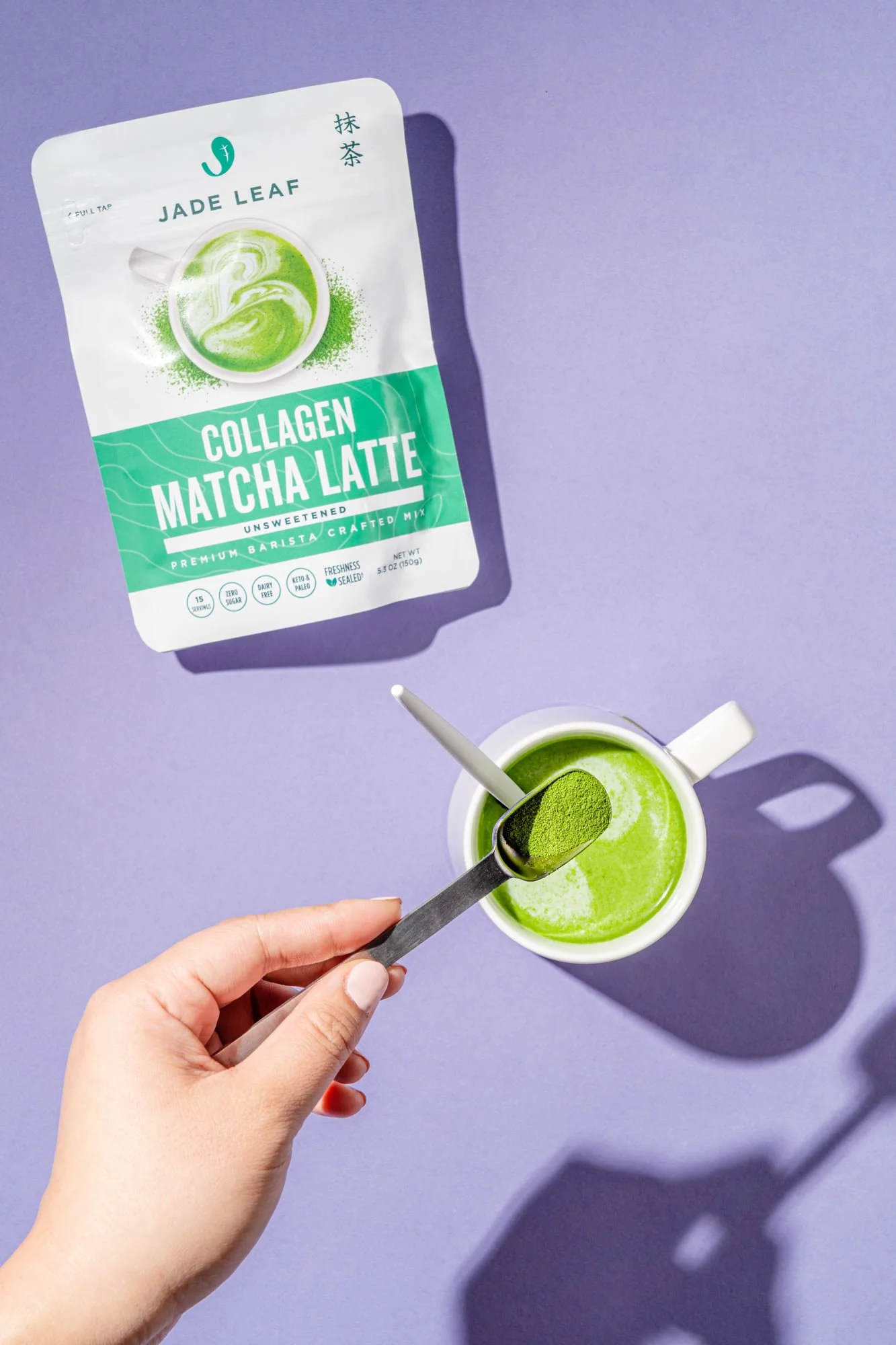 A person holding a spoon with matcha powder over a cup of green matcha latte on a purple surface, with a Jade Leaf collagen matcha latte package in the background.