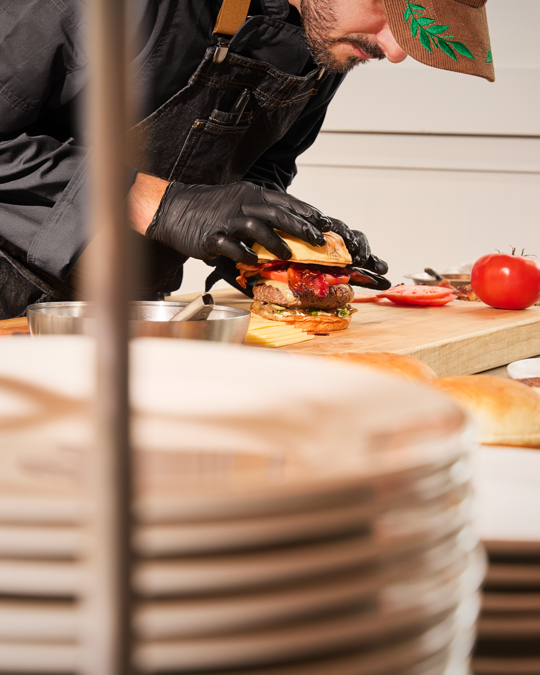 A chef wearing black gloves and a brown cap assembling a gourmet burger on a wooden cutting board, with tomatoes and condiments on the counter.