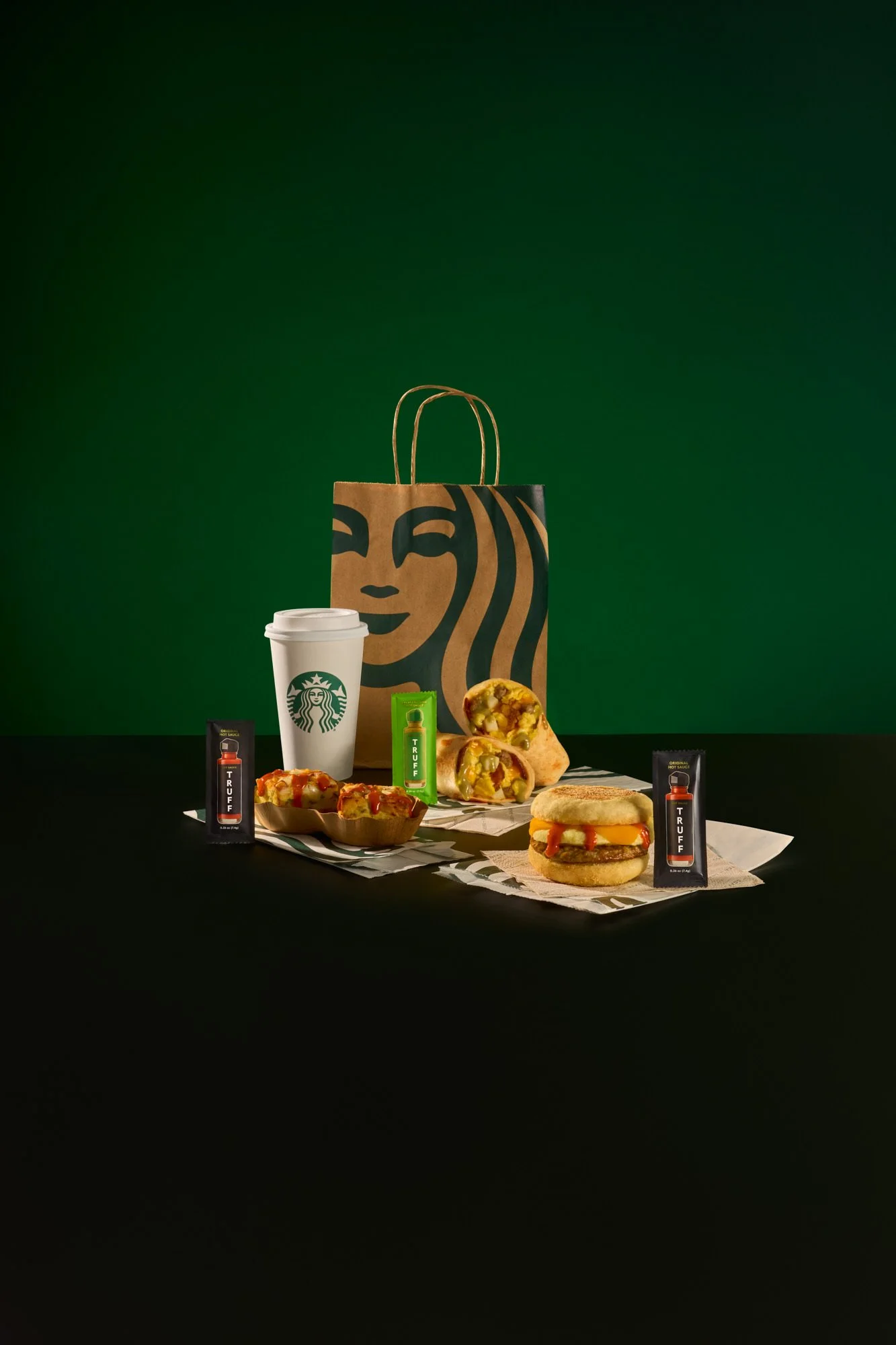 Sandwich with lettuce, bacon, barbecue sauce, and toasted bread slices on a black surface with a bottle of truffle hot sauce in the background.