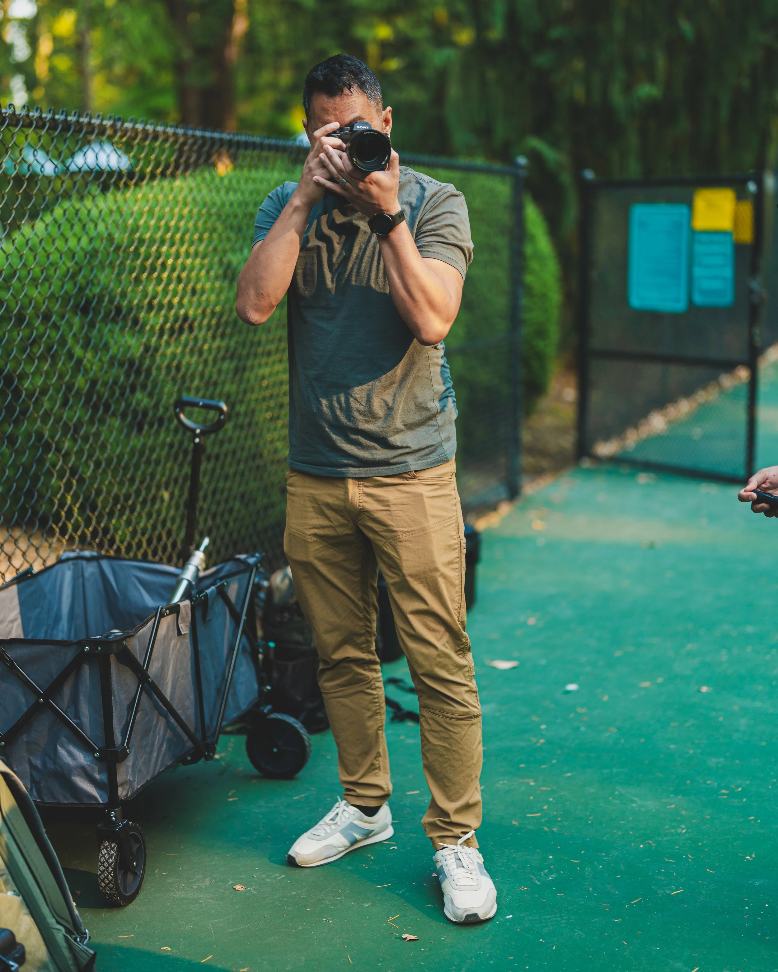 A man standing on a tennis court taking a photo with a camera, wearing a gray t-shirt, tan pants, and white sneakers, with a golf cart nearby.