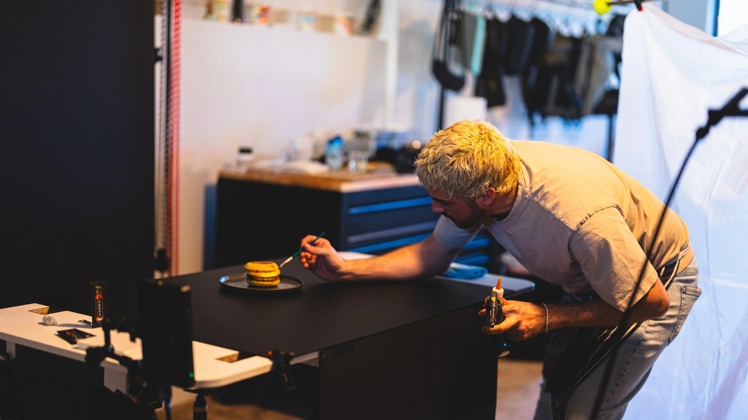 A man with blonde curly hair wearing a beige t-shirt working with a blowtorch on a small project at a black workbench in a workshop. There is a stack of macarons on a plate on the workbench, and various tools and supplies are visible around.