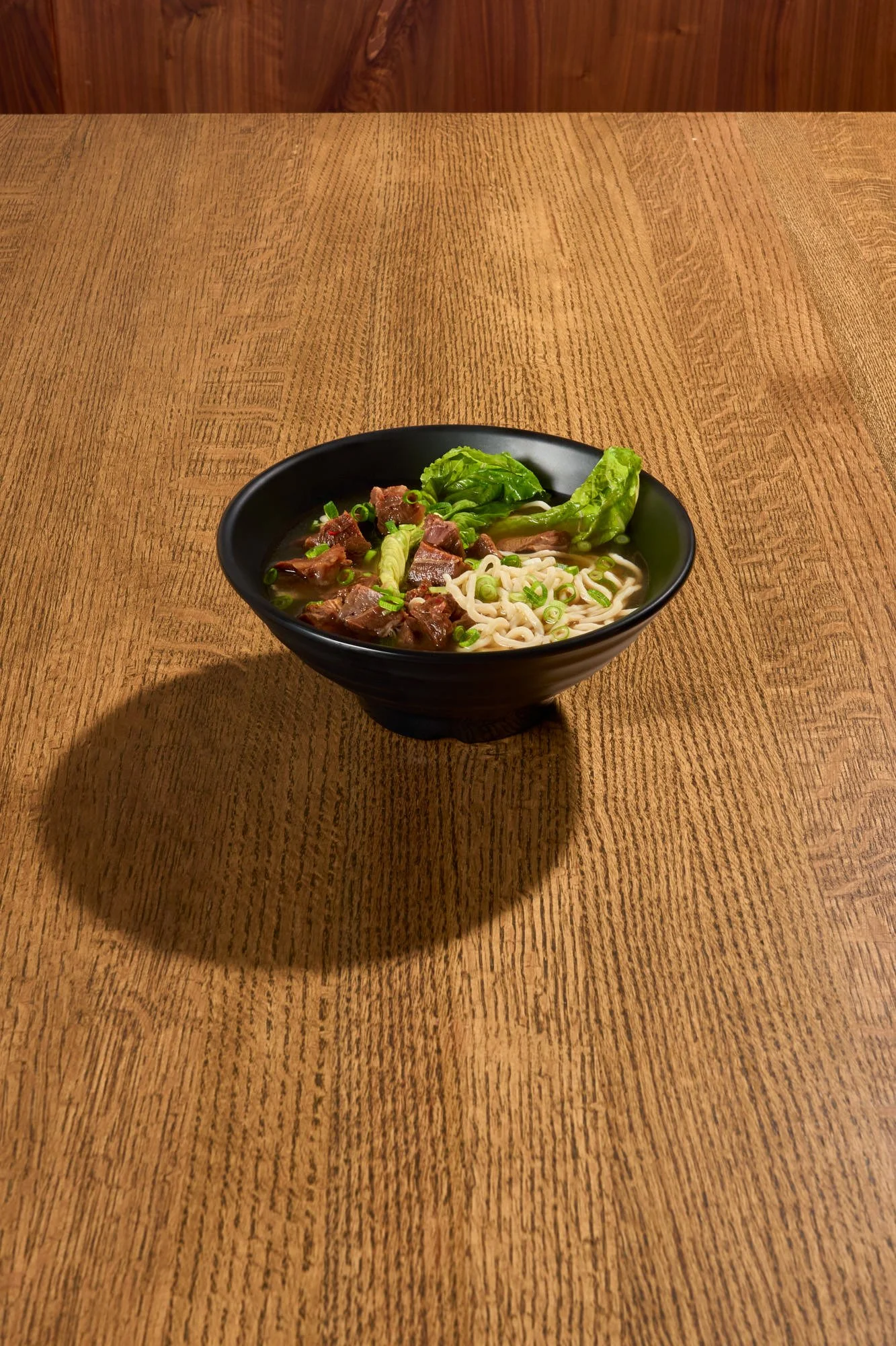 A black bowl filled with noodles, beef chunks, lettuce, and chopped green onions on a wooden table.