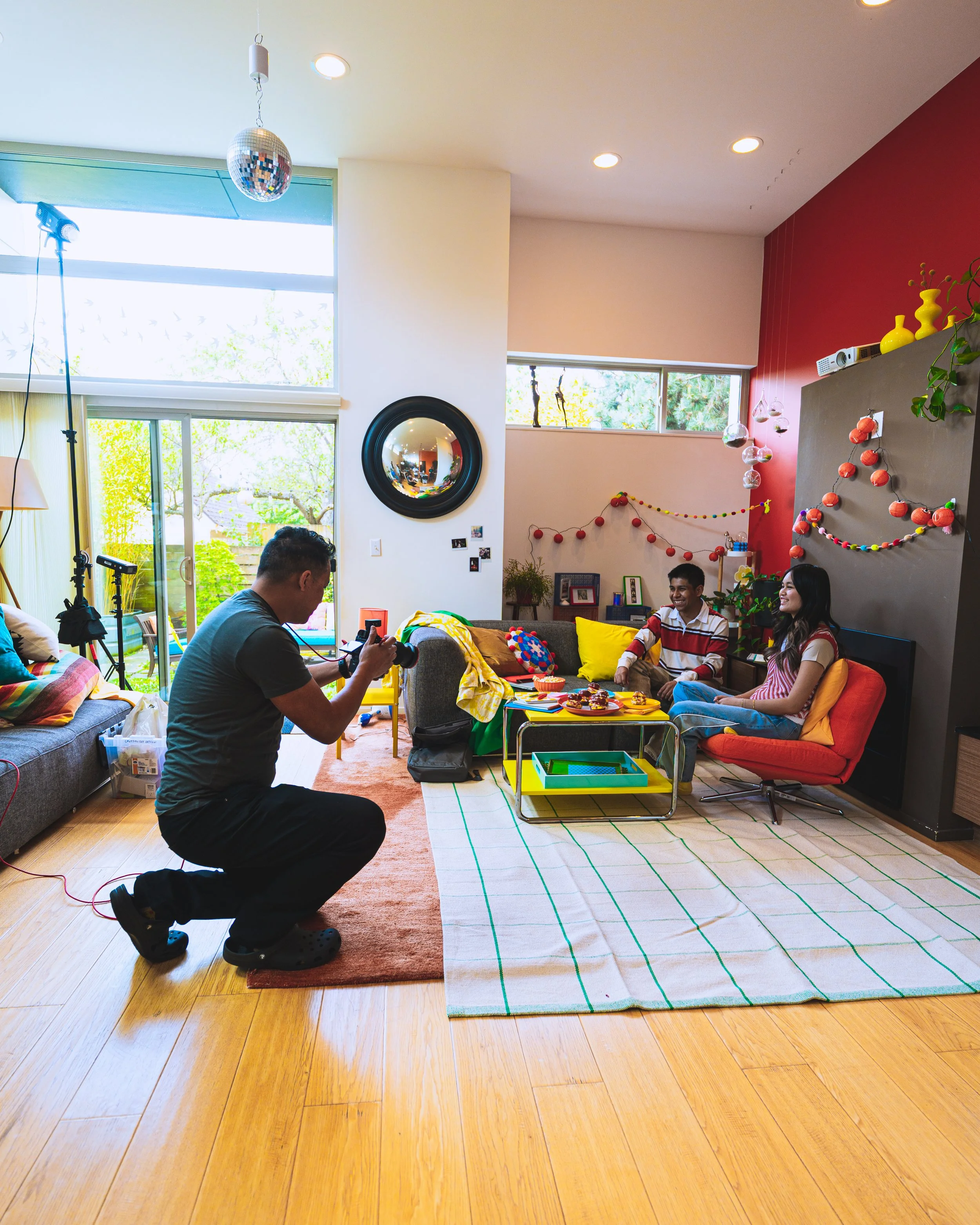 Two friends take a photo of a couple sitting on a colorful couch in a decorated living room with a large window and vibrant decor.