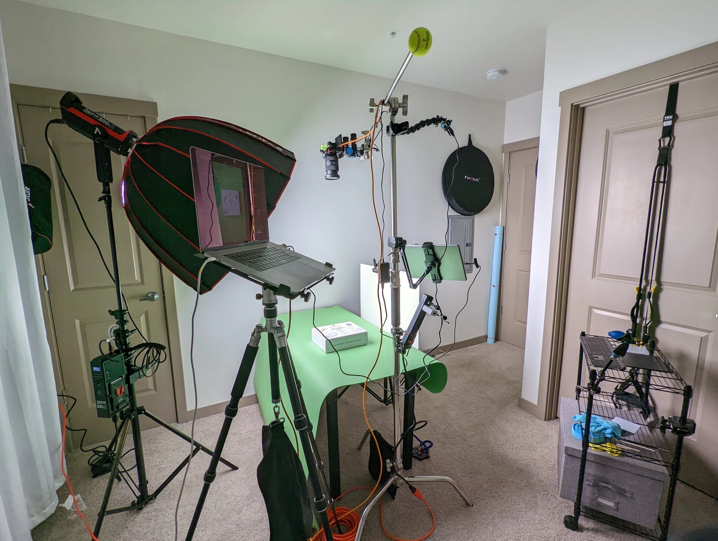 Room with photography equipment including a softbox light, a laptop on a stand, a camera mounted on a tripod, one monitor, a green screen backdrop, and other accessories for a photo or video shoot.