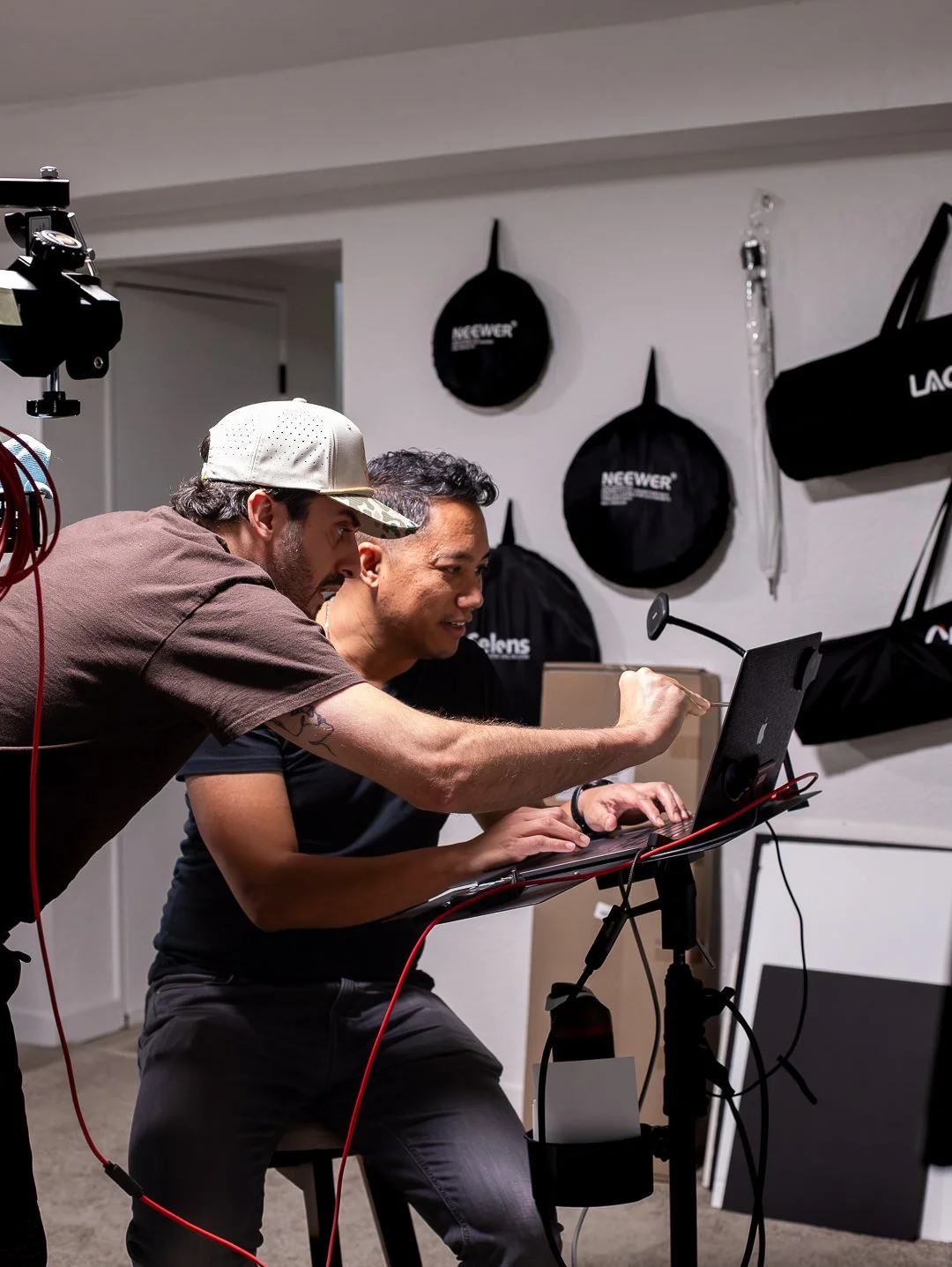 Two men working together on a computer, one sitting at a desk and one leaning over, in a room with black bags hanging on the wall.