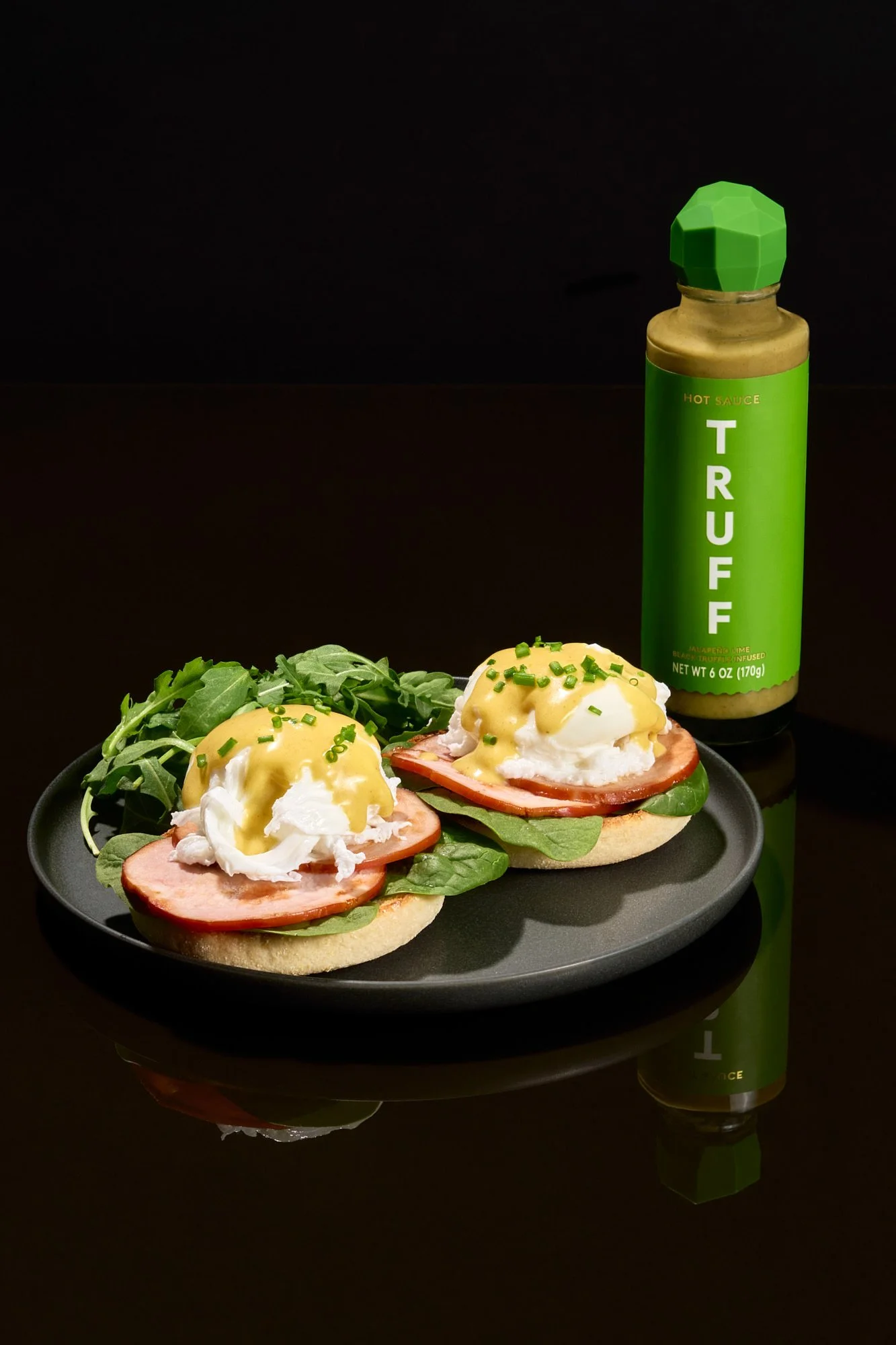 Two breakfast sandwiches with eggs, ham, lettuce, tomato, and hollandaise sauce on English muffins, served on a black plate; arugula salad and a bottle of Trader Joe's hot sauce in the background.