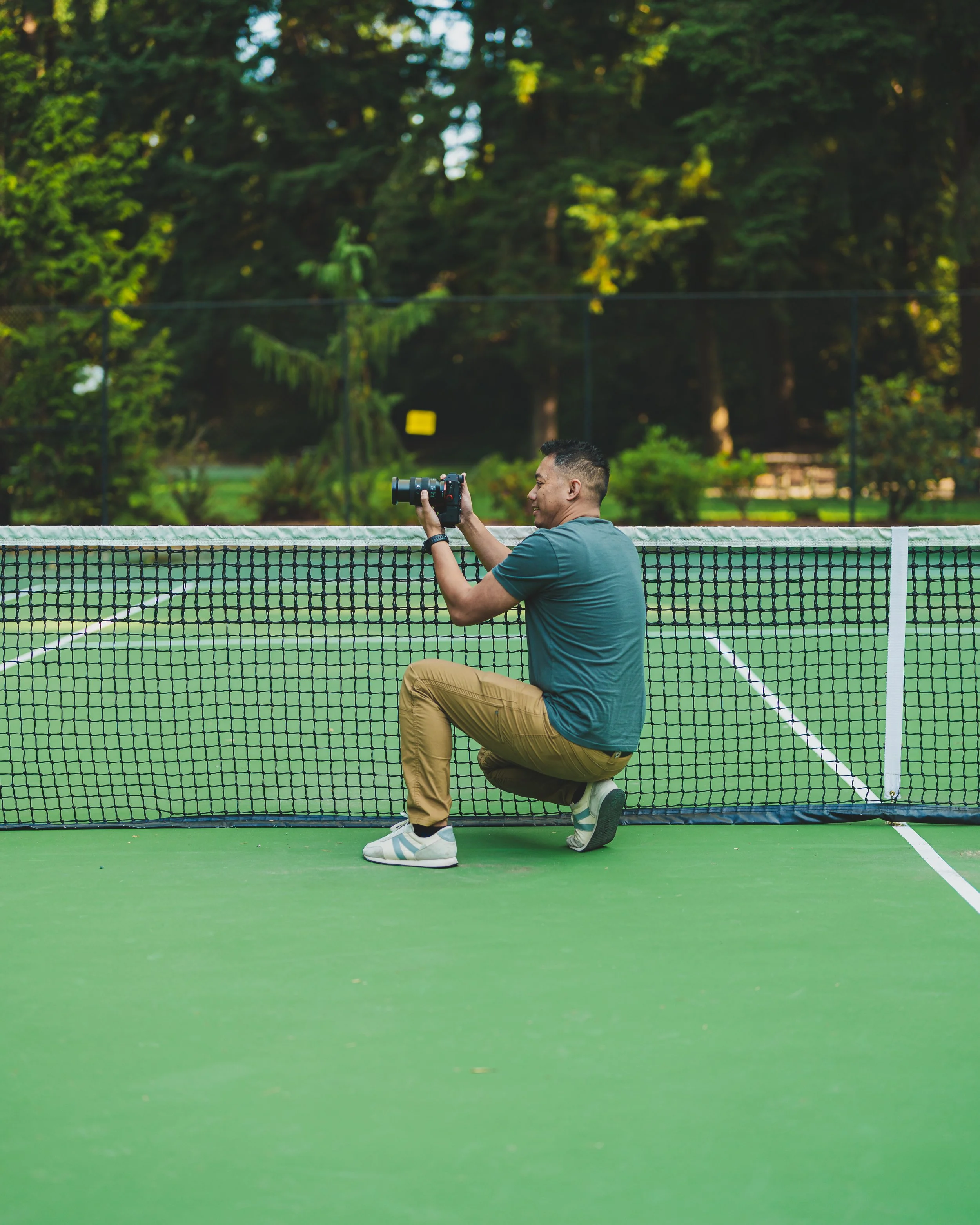 A man kneeling on a tennis court, taking a photograph through a camera lens, with a green fenced background and trees.