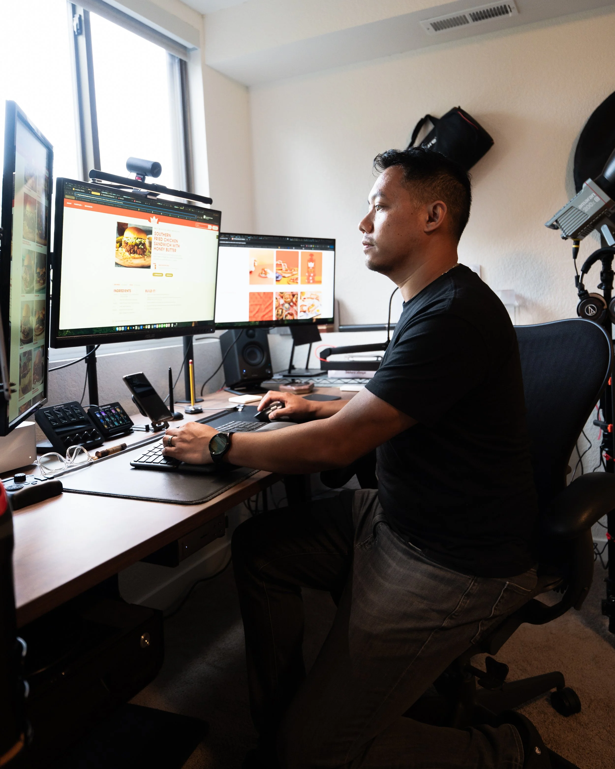 A man working at a desk with dual computer monitors, one showing a burger recipe and the other displaying food images, in a home office with natural light from a window.