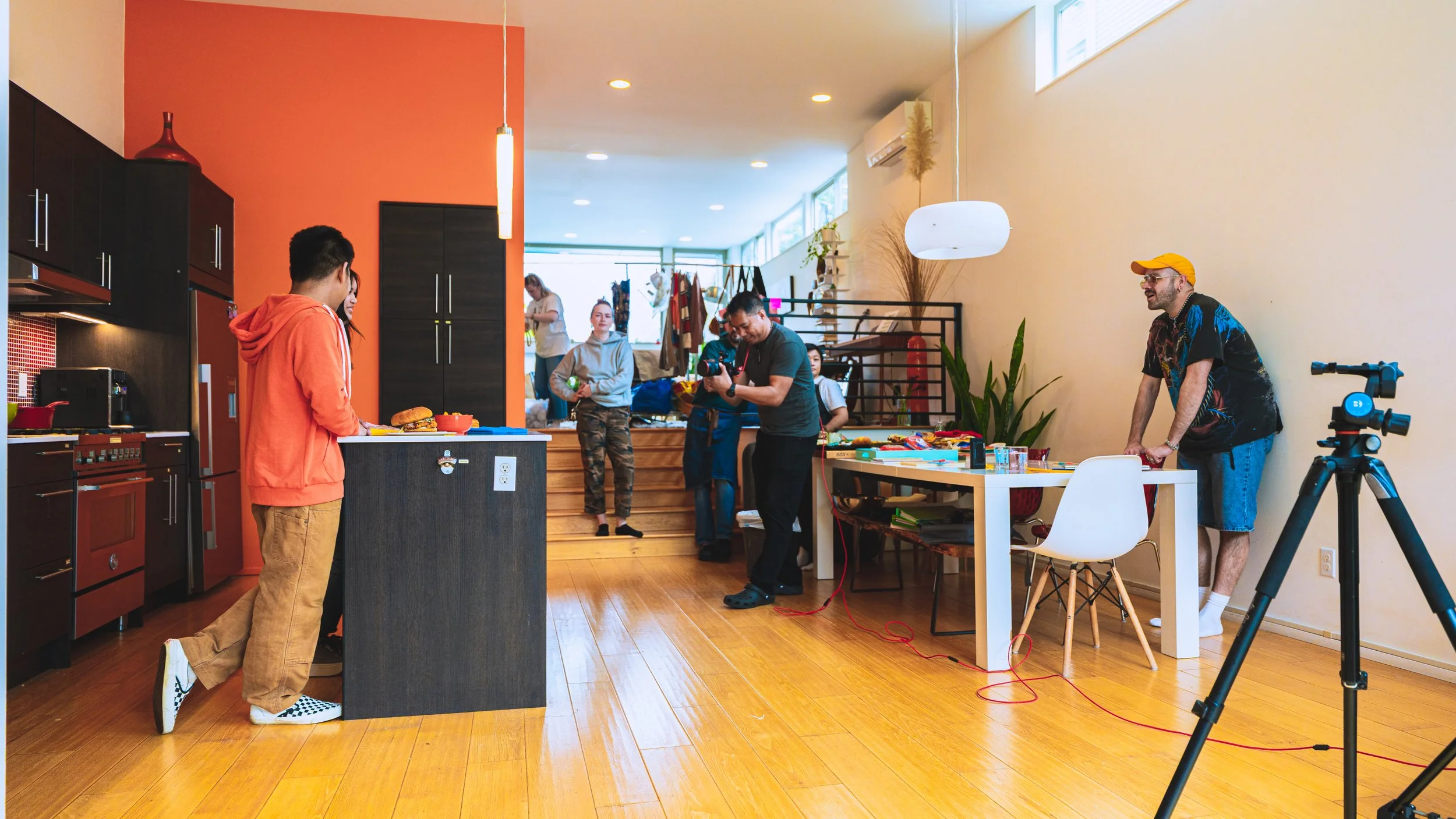 People in a modern home studio setting, preparing for a photo or video shoot, with cameras, lighting, and varied activities happening around.