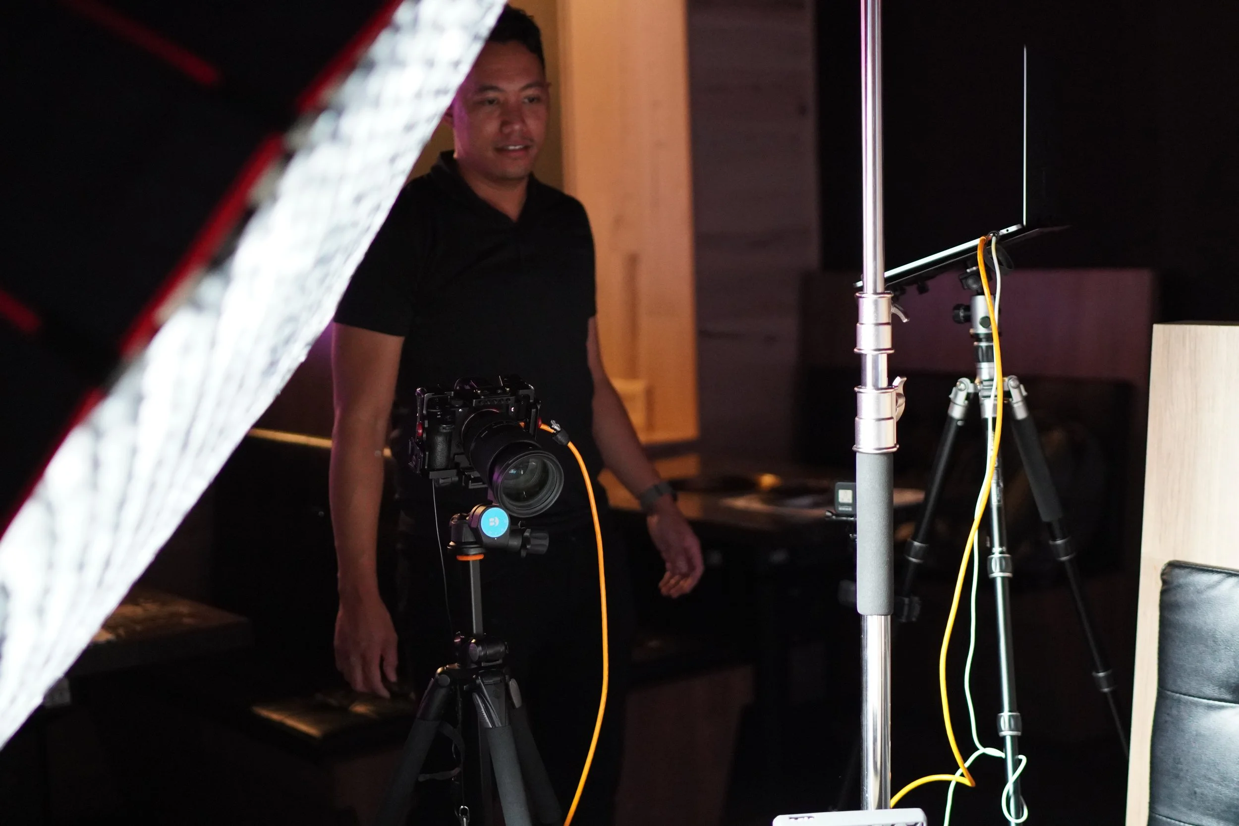 A man standing in a dimly lit room with professional camera and lighting equipment, looking towards the camera.