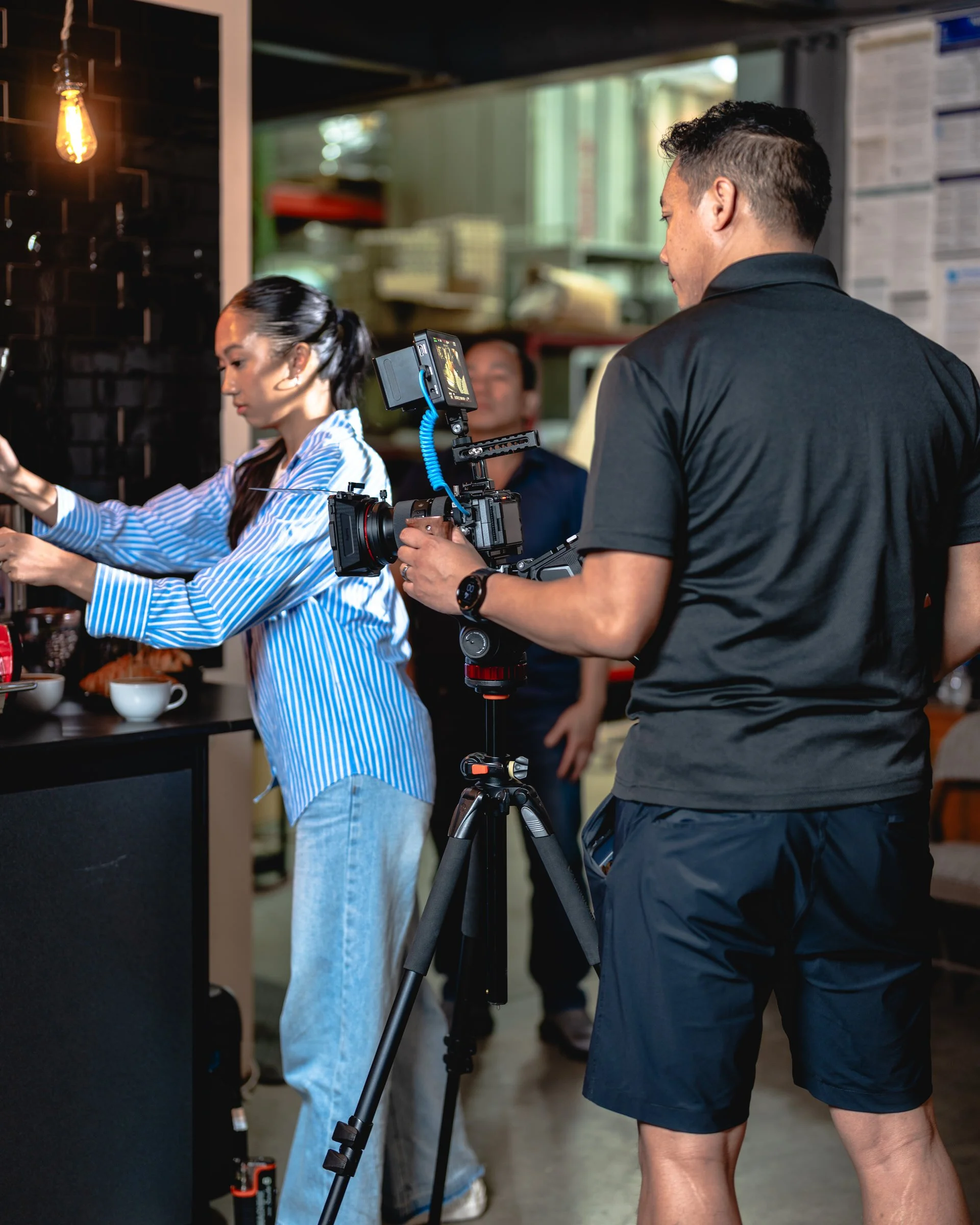 Filming inside a restaurant or cafe, a man in black shirt and shorts is holding a camera on a tripod, filming a woman in a blue and white striped shirt preparing food or drinks behind the counter.