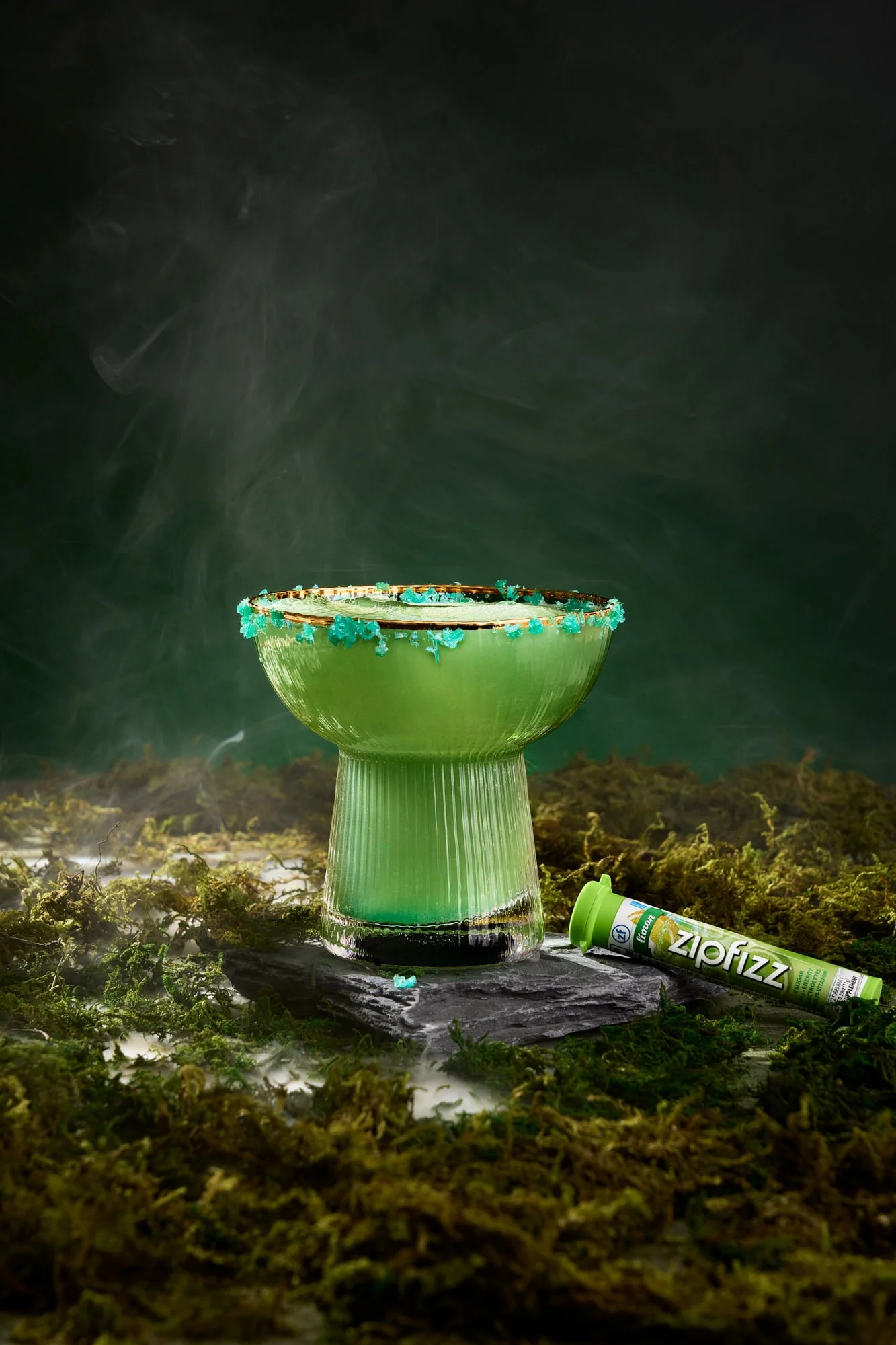 A green glass cup emitting blue smoke, with a tube labeled "Zipfizz" lying on mossy ground, in an outdoor scene with a dark, smoky background.