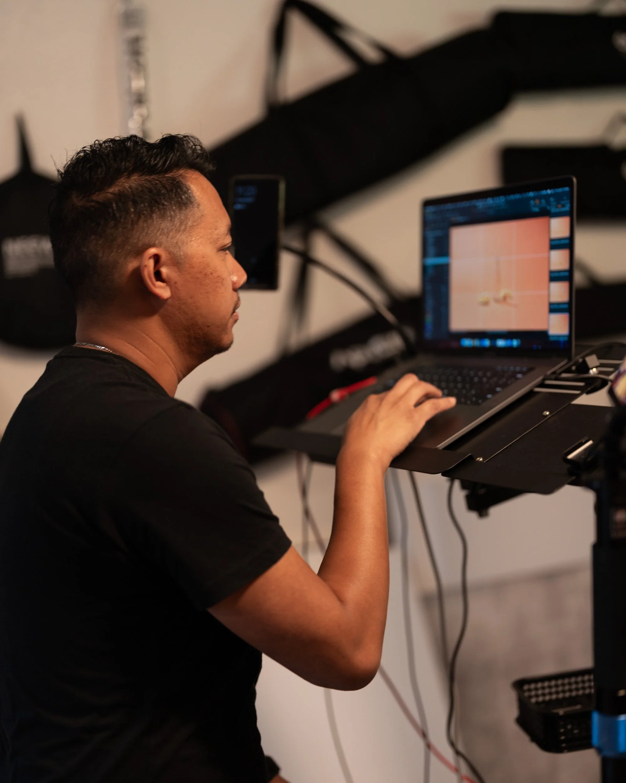 A man with short black hair working on a laptop with a pink and orange screen, sitting at a desk with a keyboard and some electronic equipment. His left arm is resting on the desk, and he is looking at the laptop screen.