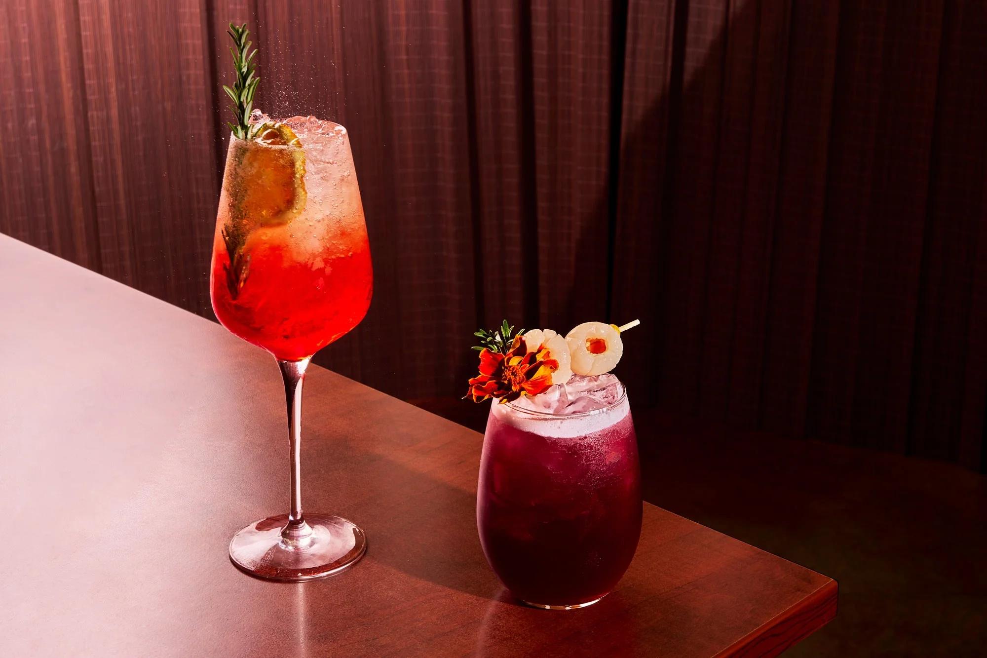 A tall glass of red cocktail garnished with a slice of lemon and a sprig of rosemary, next to two shorter glasses of pink drinks topped with fruit and herbs, on a wooden bar or table with a dark wood background.