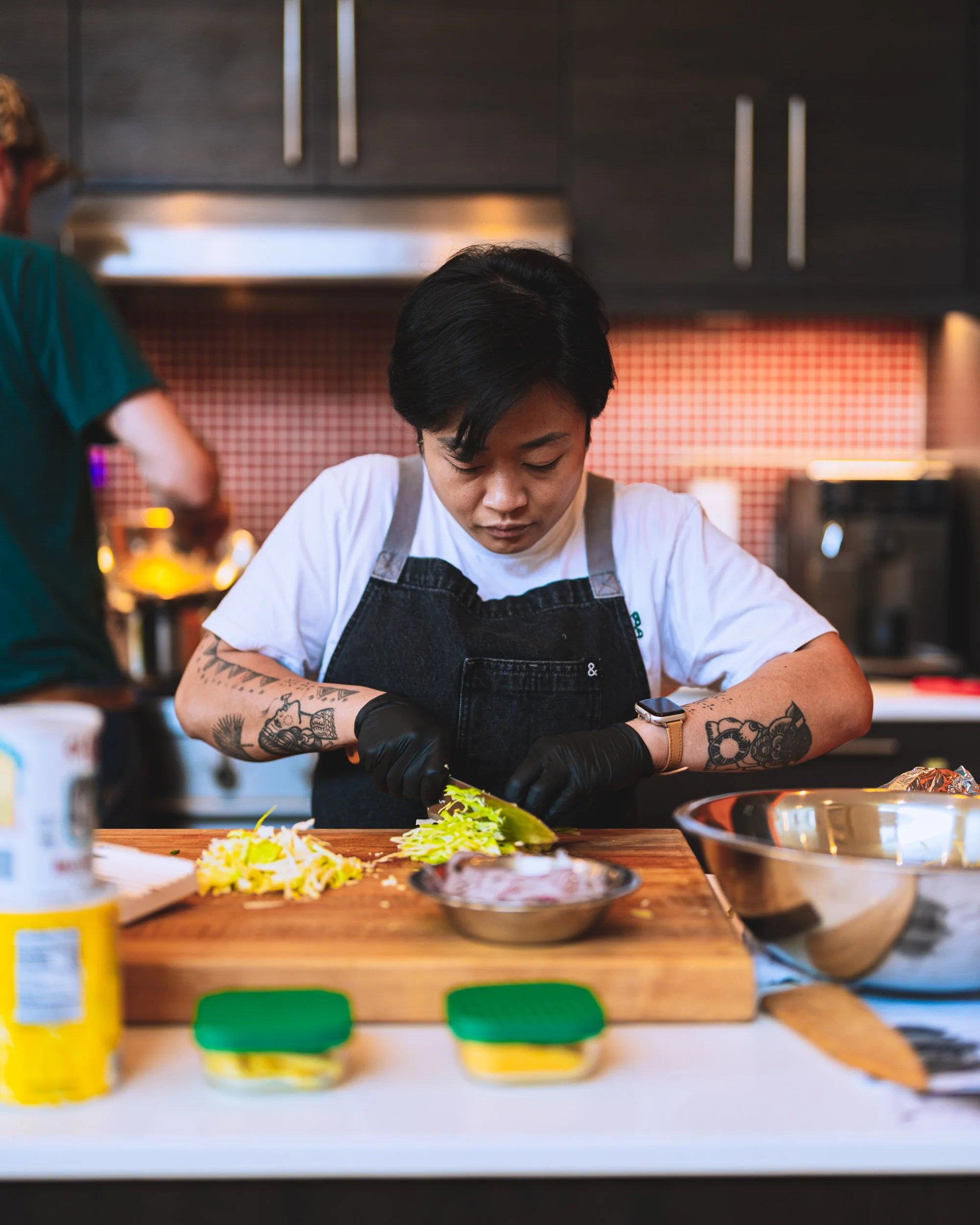 A person with dark hair, tattoos on their arms, wearing a white shirt, black gloves, and a smartwatch, is preparing food in a kitchen. They are chopping yellow vegetables on a wooden cutting board with various bowls and ingredients around.