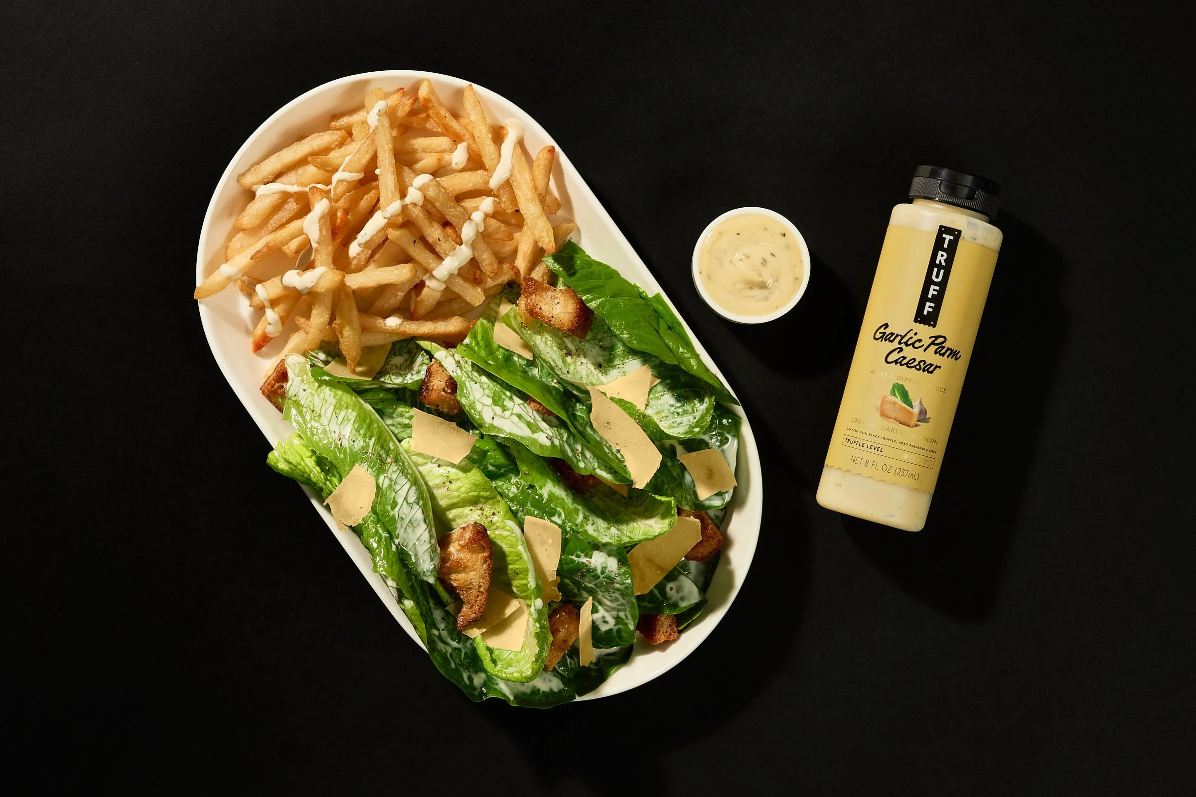 A plate of French fries with a drizzle of white sauce, a small cup of creamy dressing, and a salad with romaine lettuce, shaved cheese, and croutons. Next to the plate is a bottle of garlic Parmesan Caesar dressing.
