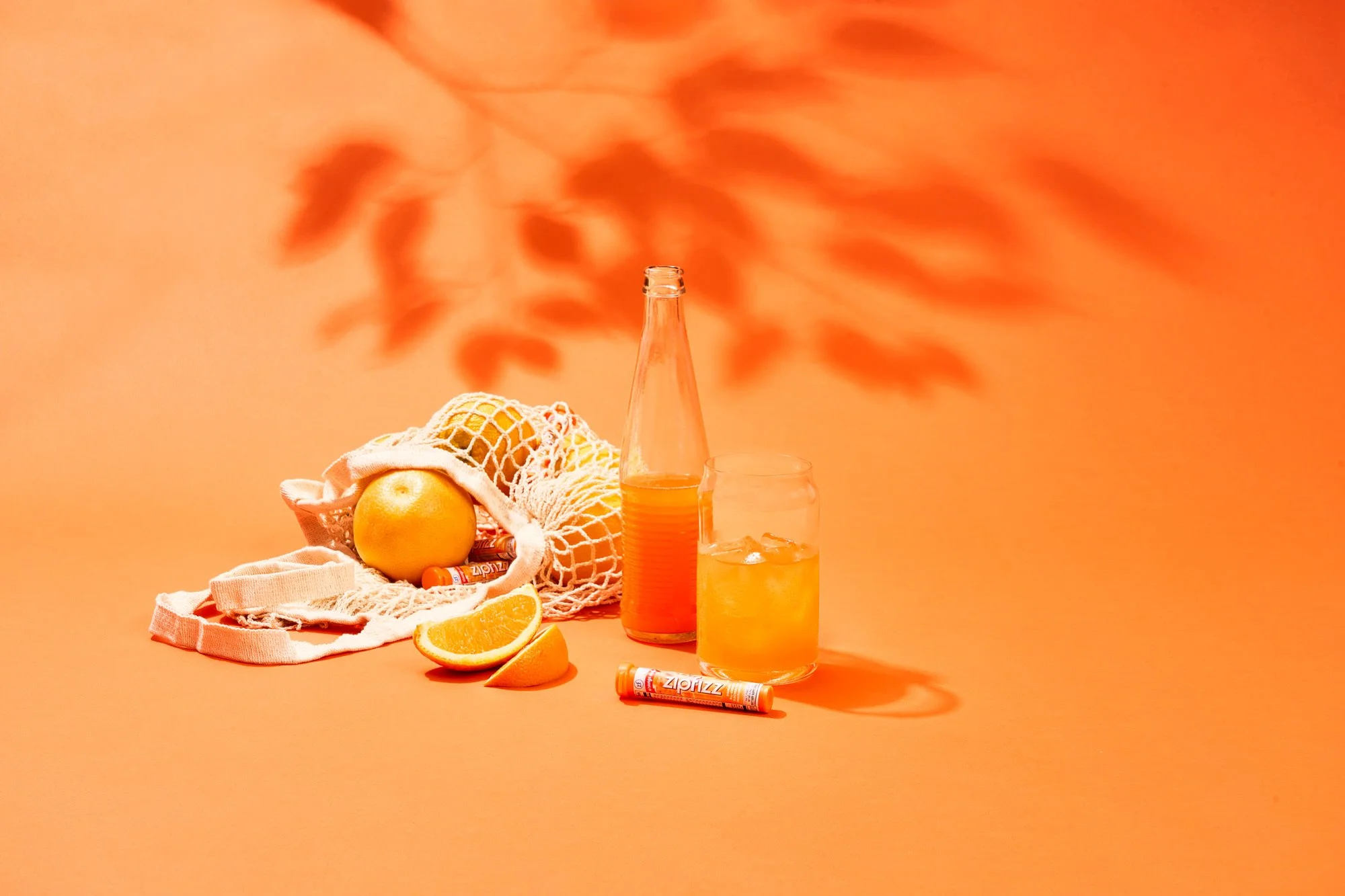 A still life composition with oranges, sliced orange wedges, a glass of orange juice with ice, a bottle of orange soda, and a mesh bag of oranges on a peach-colored background.