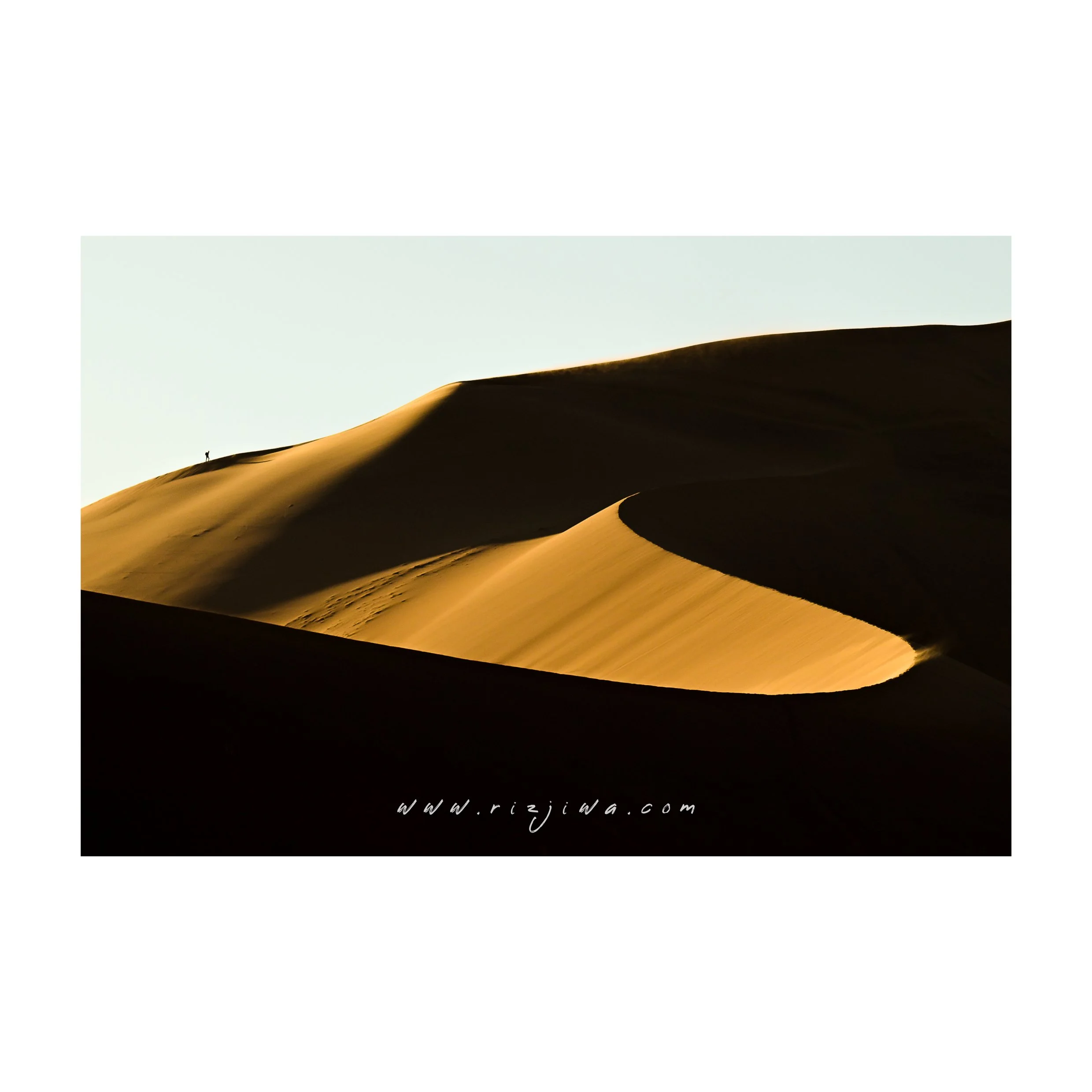 Sand dunes with a small human figure walking on top, visible against a clear sky.