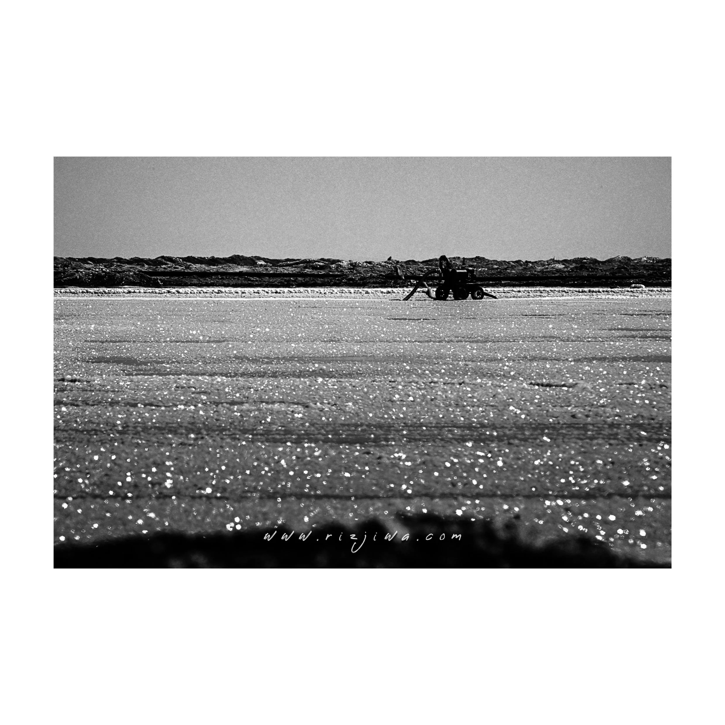 Black and white photo of a sandy beach with a small vehicle and someone leaning on it, in the distance along the shoreline.