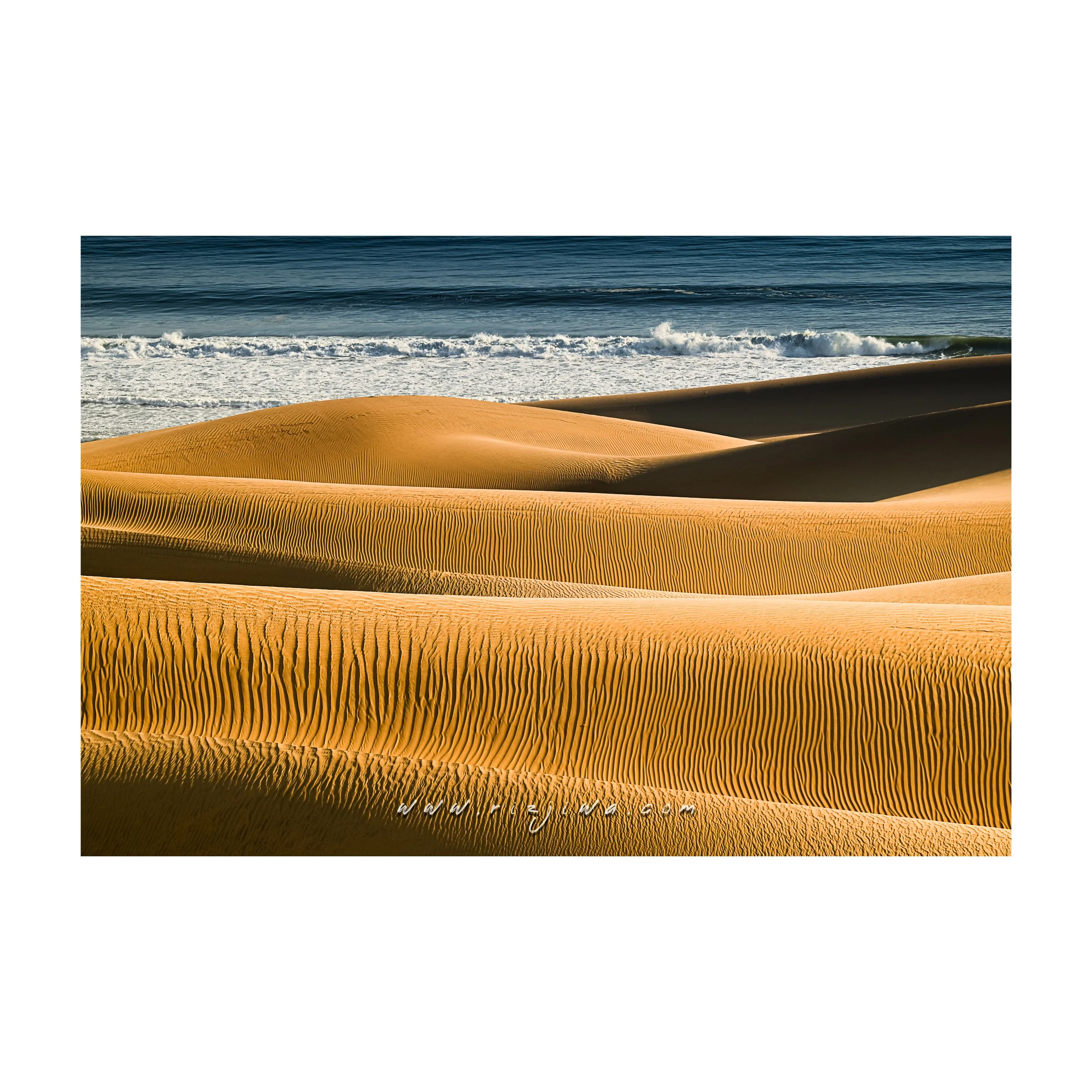 Photo of golden sand dunes with rippled patterns along the beach, ocean waves in the background under a clear sky.